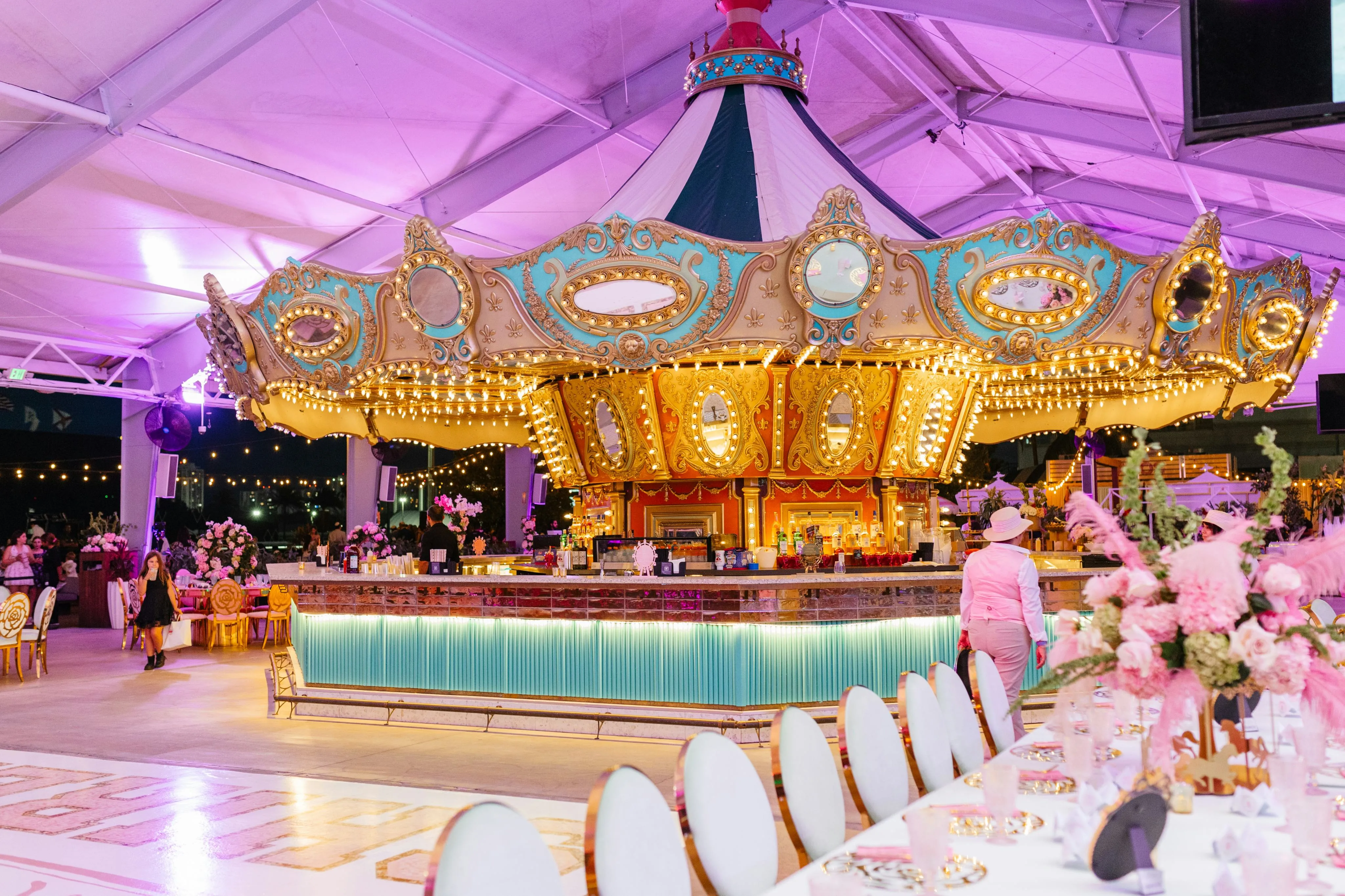 Ornate carousel-themed bar lit with warm lights in an elegant event space with floral decorations and dining tables.