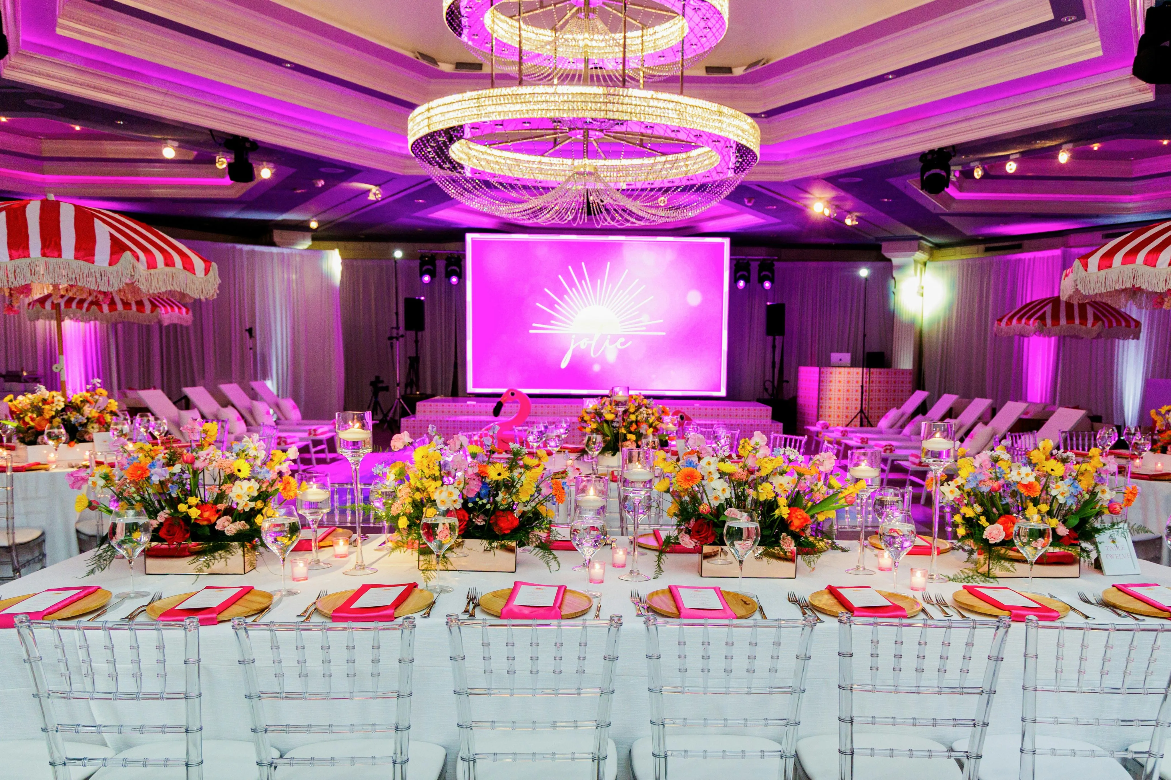 Elegant banquet hall with white tables set with colorful floral centerpieces, glassware, gold chargers, and pink napkins under a large crystal chandelier with purple lighting.