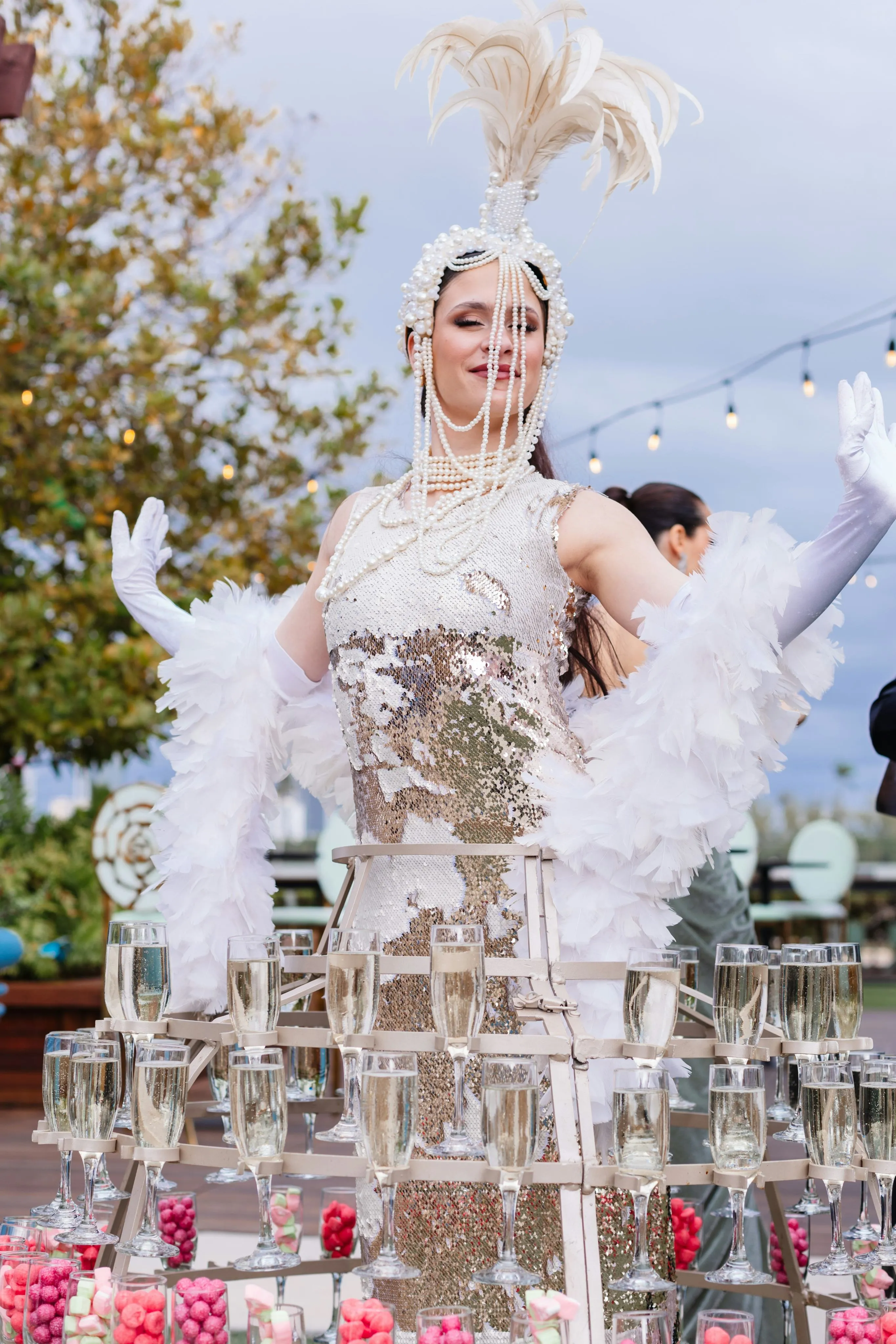 Woman dressed in a sequined gown with a feathered headpiece and pearl decorations, standing behind a circular frame holding multiple champagne glasses.