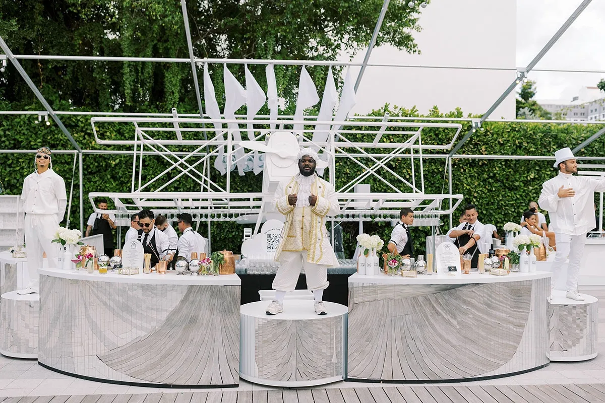 Man dressed in white and gold standing on a mirrored pedestal at an elegant outdoor bar with bartenders and white floral arrangements.