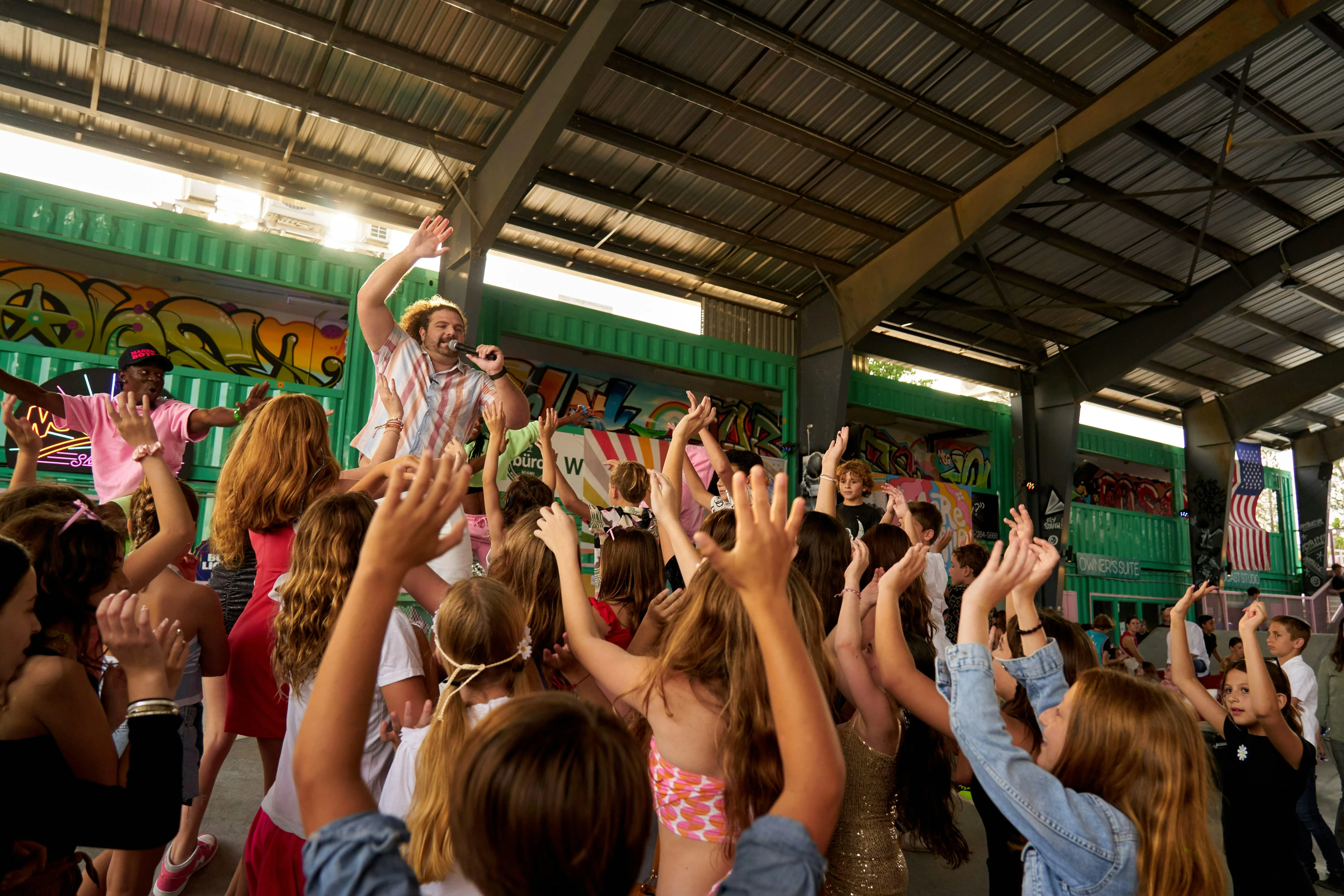 A man with a microphone leads a crowd of children raising their hands at an indoor event with colorful graffiti on walls.