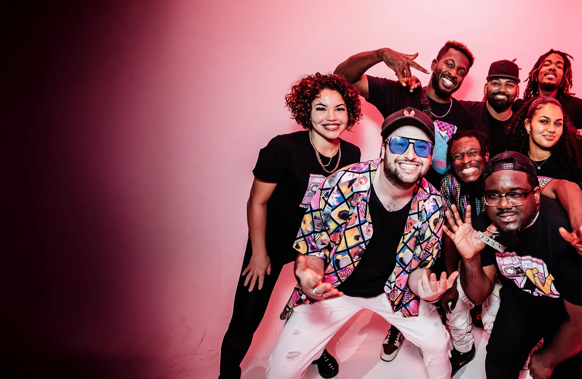 Group of eight diverse young adults smiling and posing enthusiastically against a pink background.
