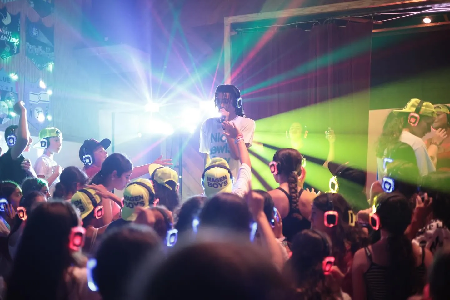 Crowd wearing glowing headphones watching a performer singing with colorful lights in the background at a silent disco event.