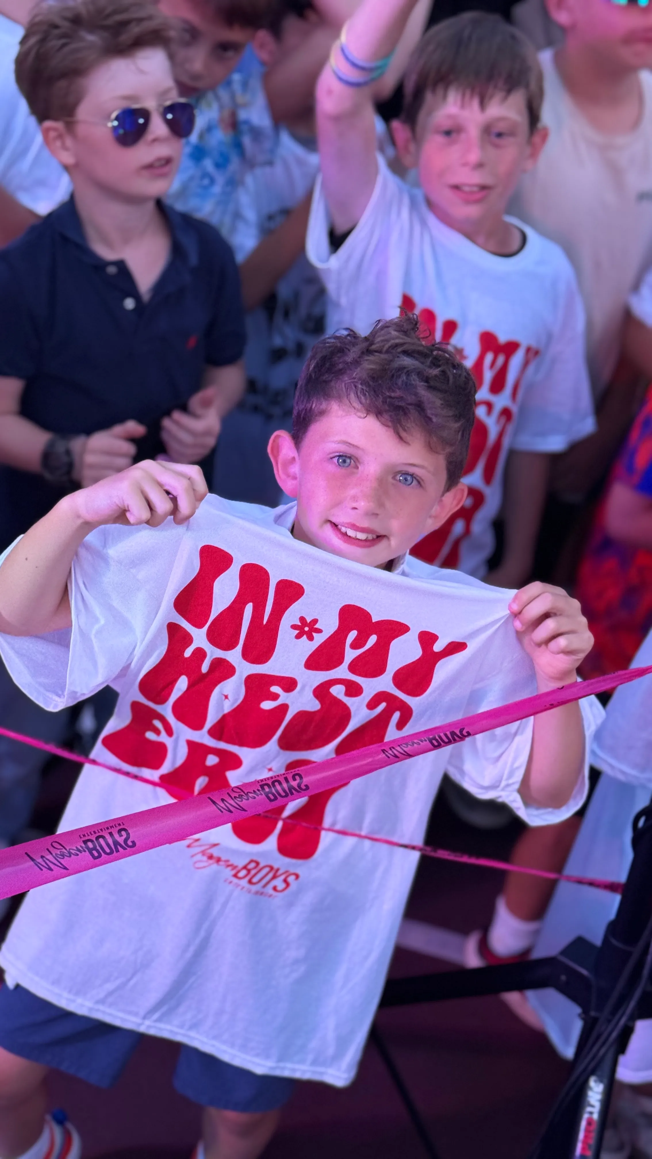 Smiling boy with blue eyes holding up a white T-shirt with red text saying 'IN MY WEST ERN' in a crowd at an event.