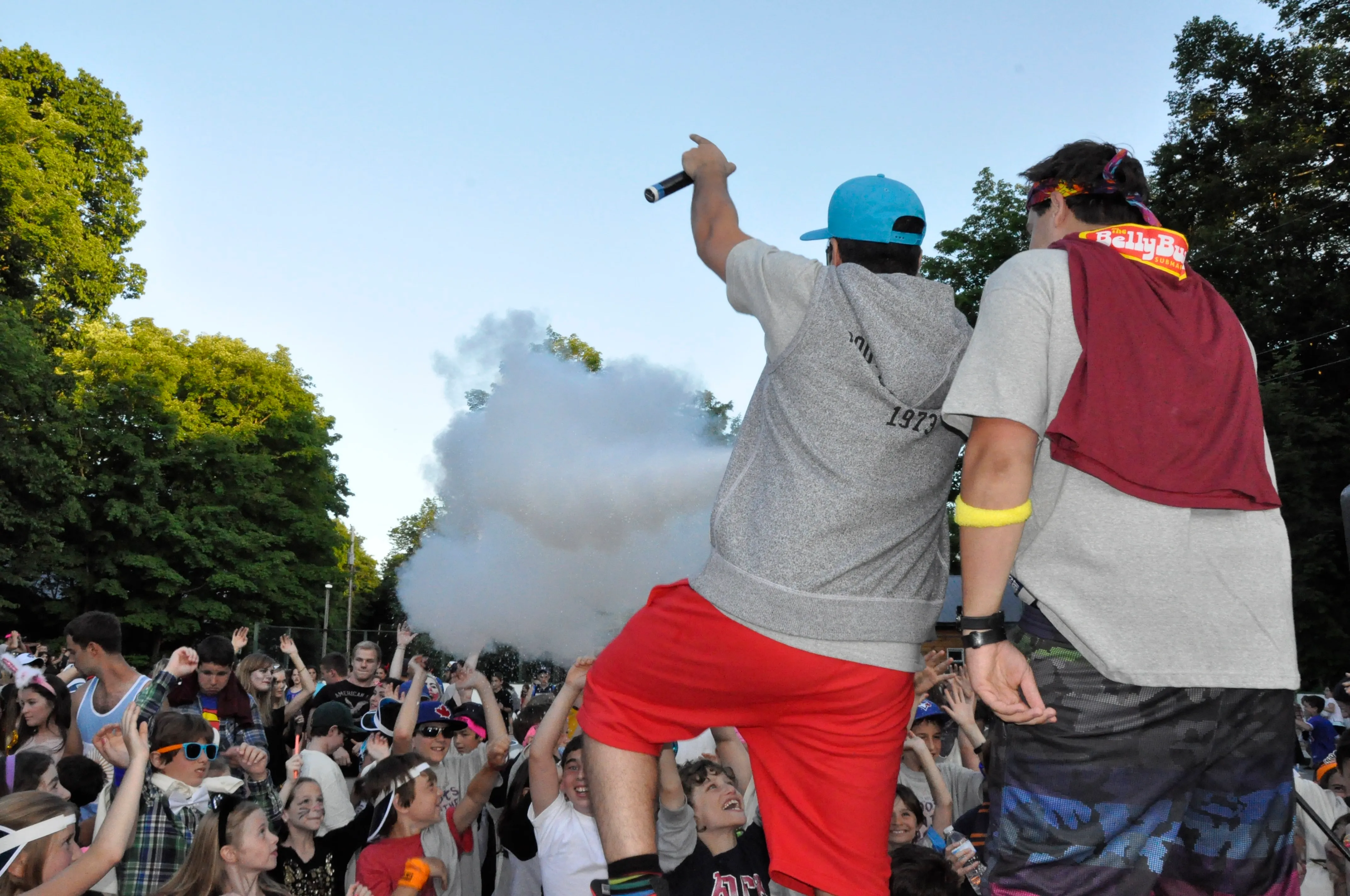 Two men facing a crowd, one holding a microphone with smoke or fog in the air, and an enthusiastic audience with raised hands outdoors.