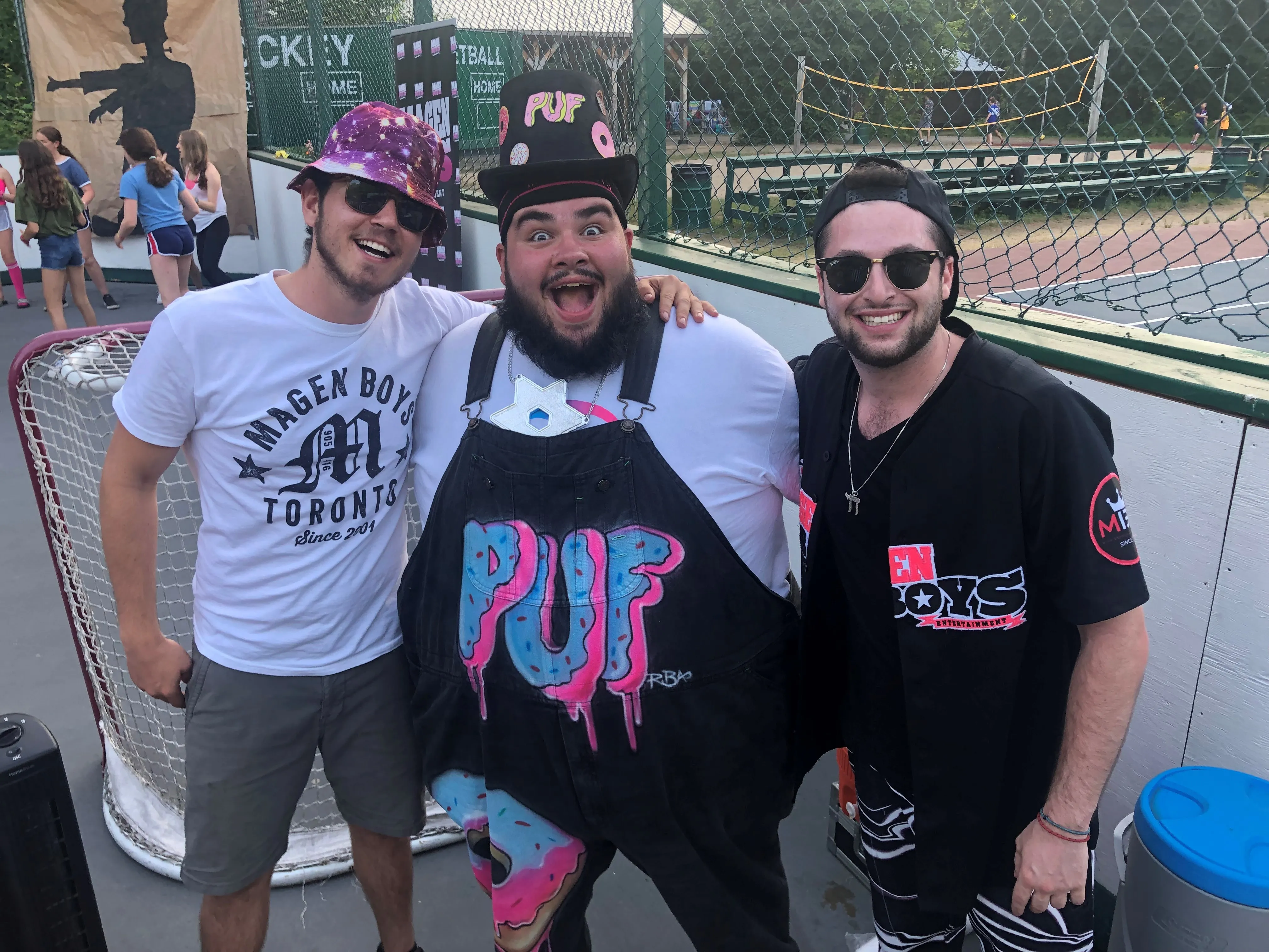 Three smiling men wearing sunglasses posing together on an outdoor sports court near a hockey net.