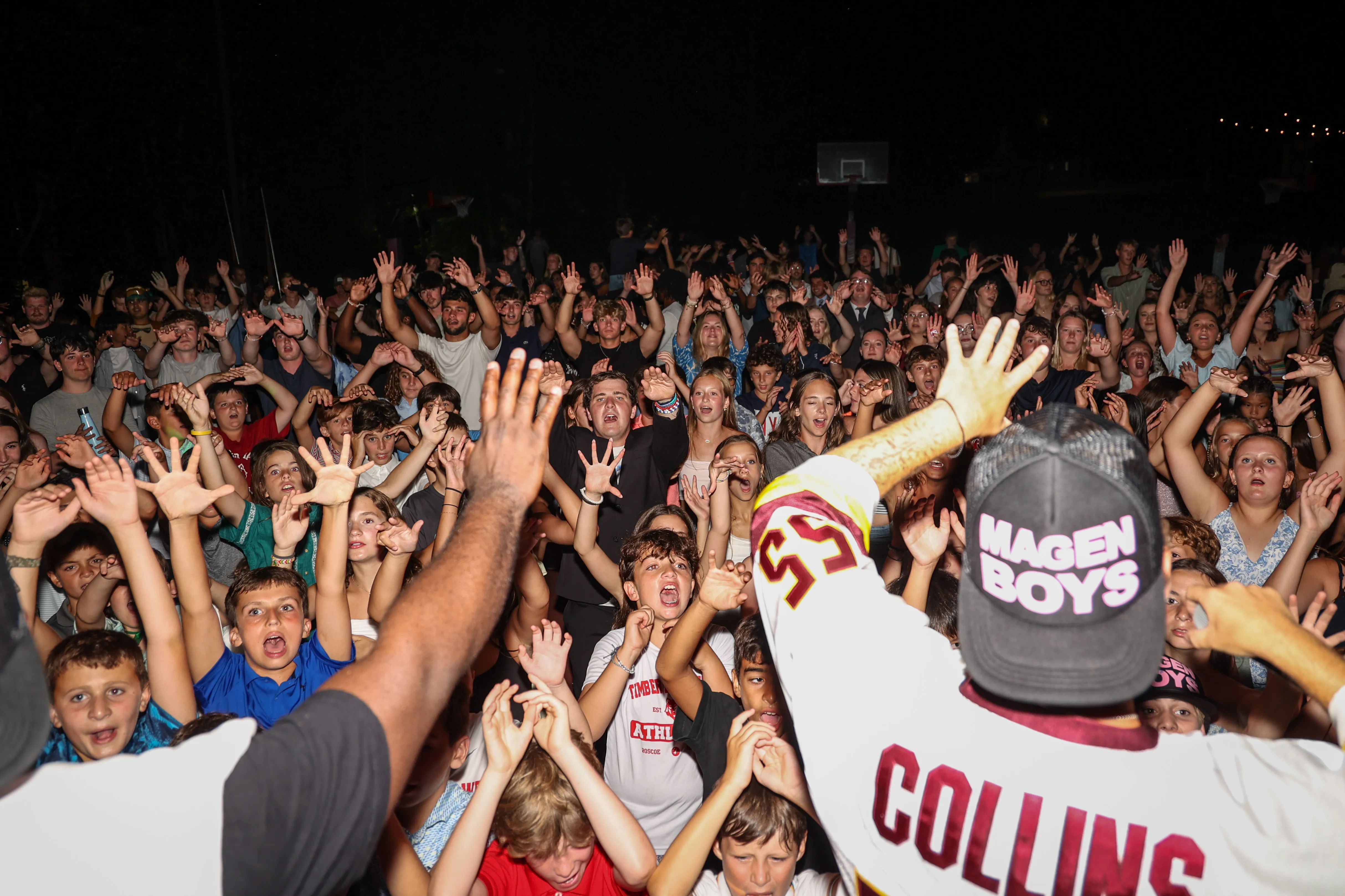 Crowd of excited young people at a night concert raising their hands toward a performer wearing a 'COLLINS' jersey and a 'MAGEN BOYS' cap.