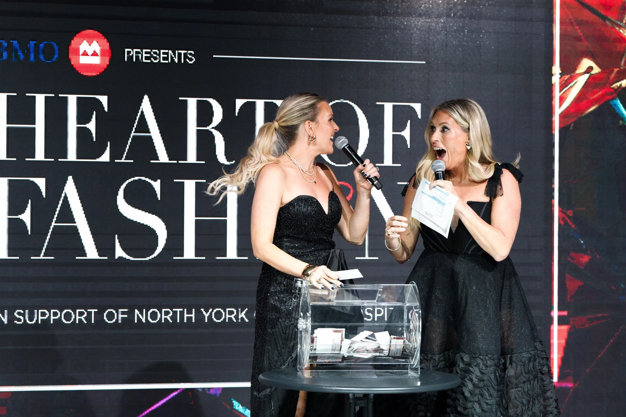Two women in black dresses hosting a raffle drawing on stage with microphones and a transparent raffle drum filled with tickets.