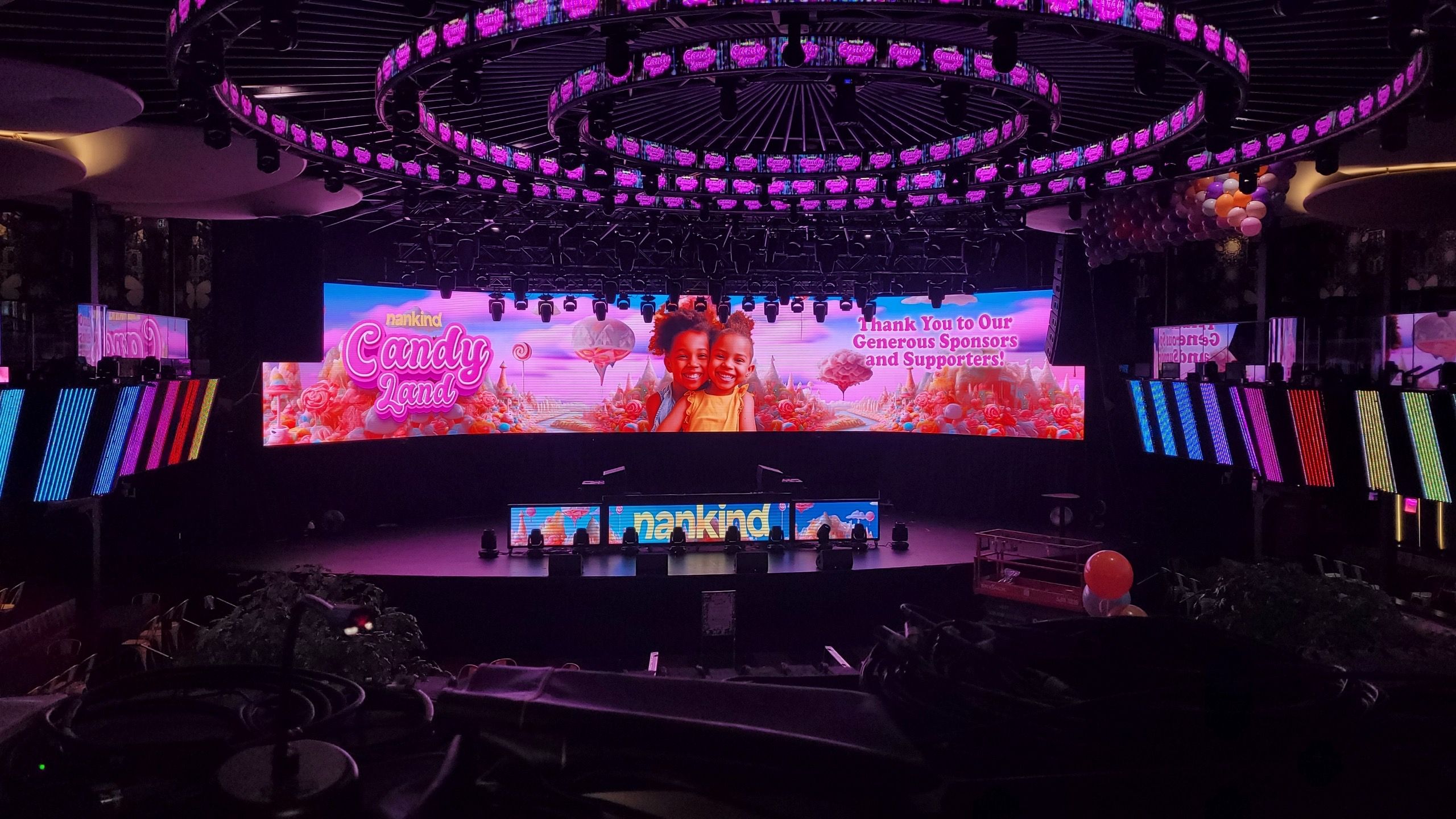Indoor stage setup with colorful LED screens displaying 'nankind Candy Land' and two smiling children, surrounded by candy-themed graphics and pink lighting.