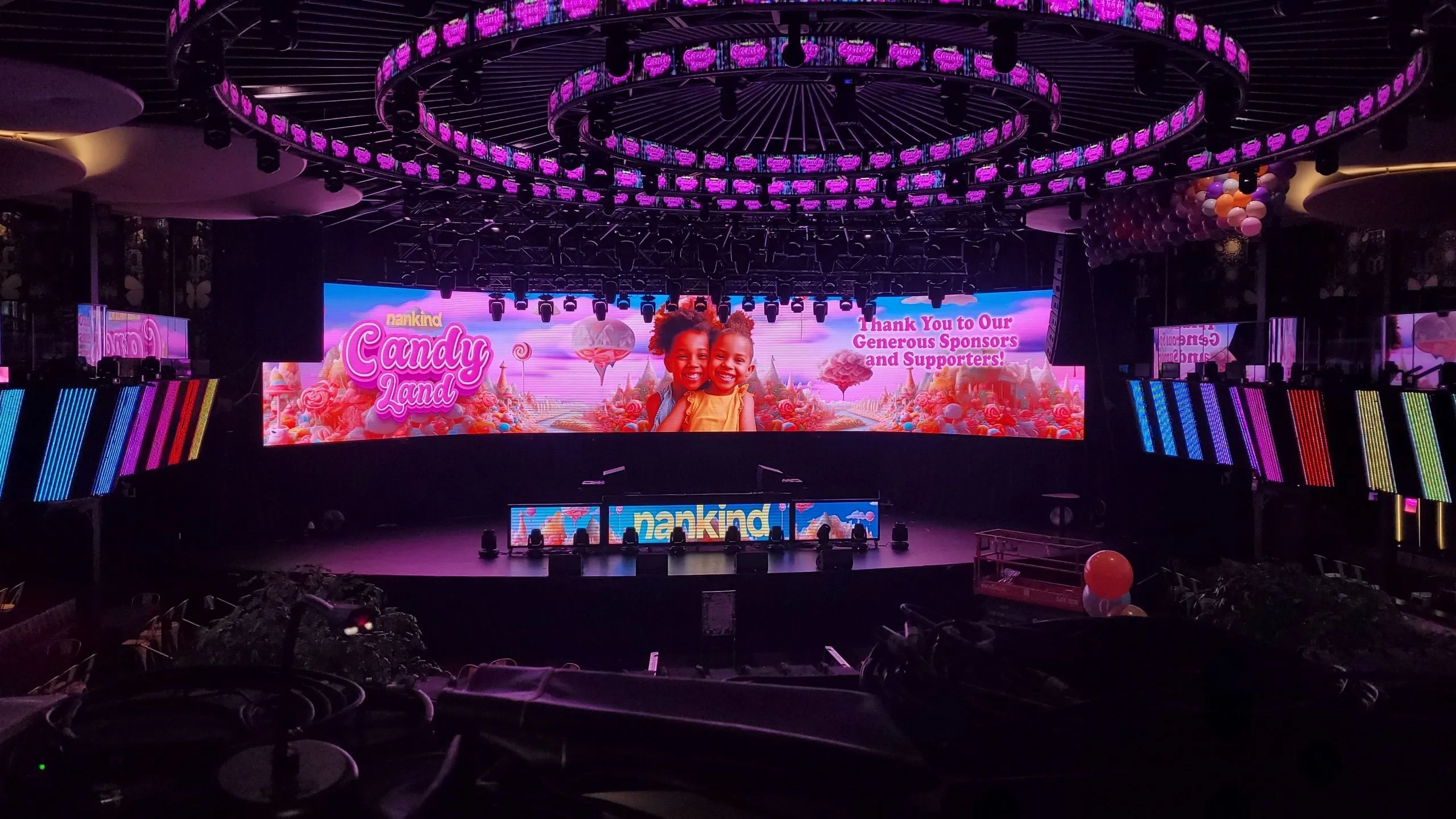 Indoor stage setup with colorful LED screens displaying 'nankind Candy Land' and two smiling children, surrounded by candy-themed graphics and pink lighting.