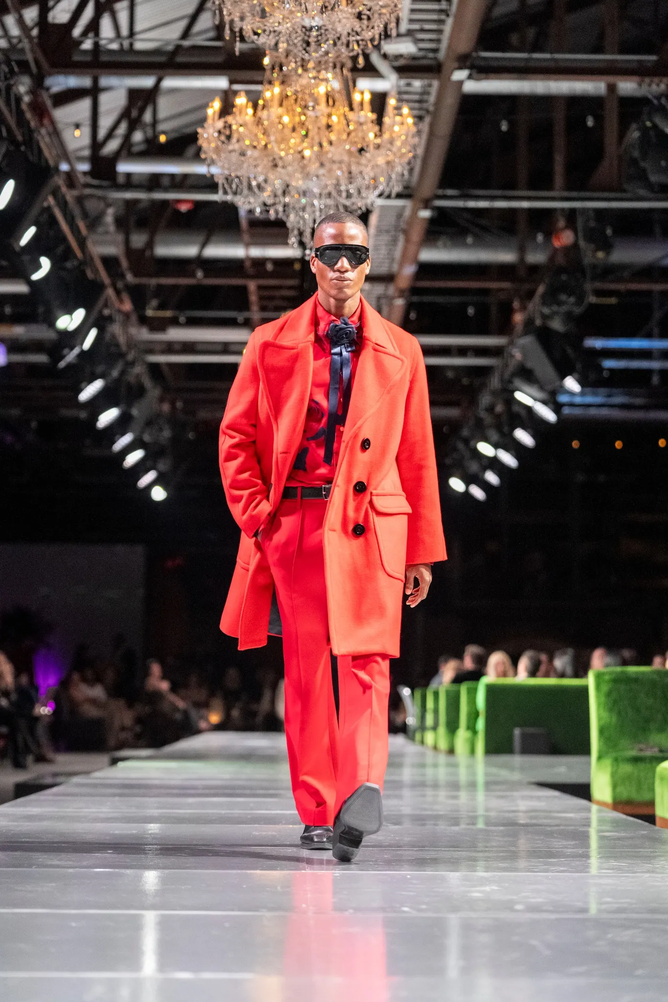 Male model wearing bright red suit and coat with black sunglasses walking down a runway under a chandelier.