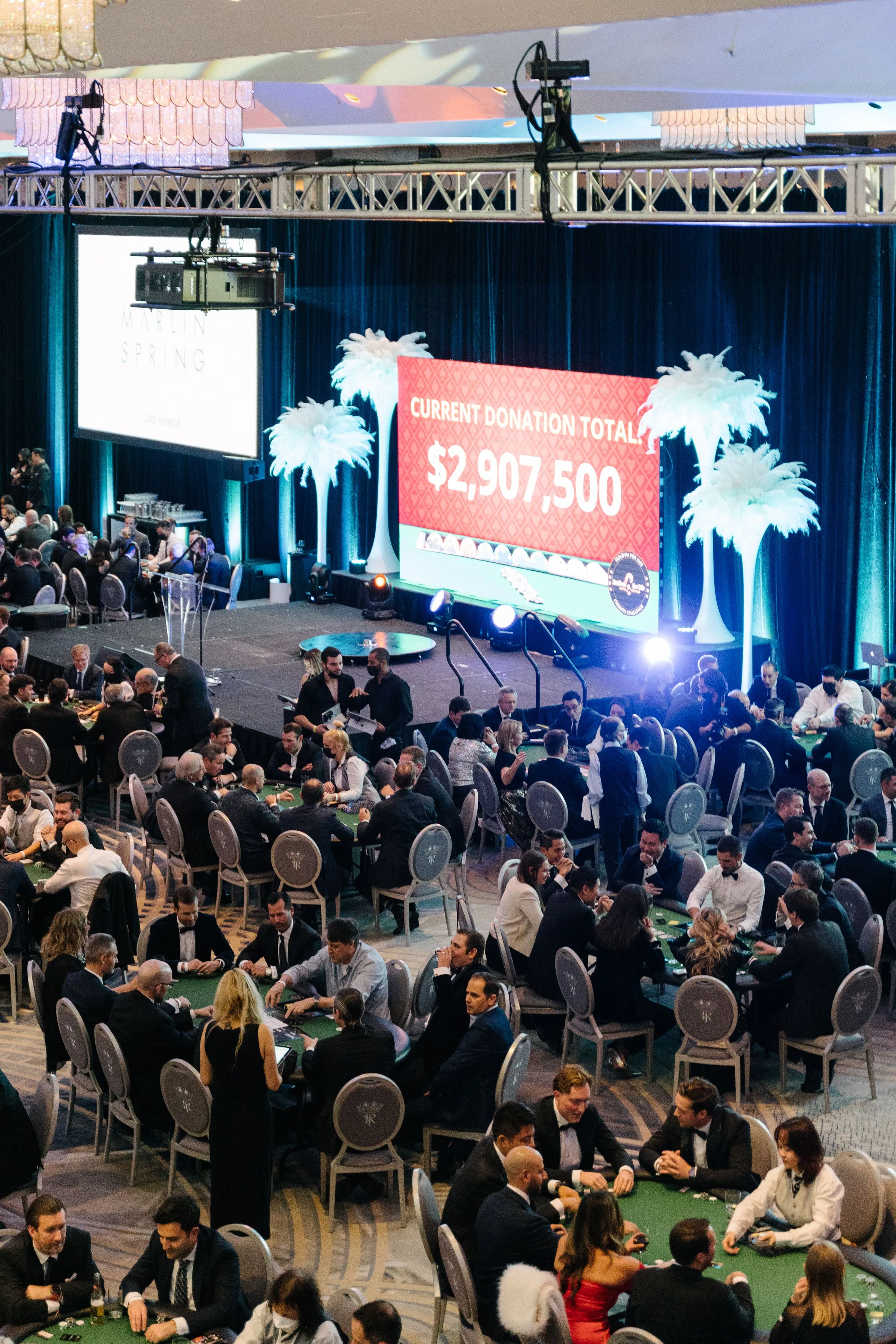 Large formal event with many people seated at green poker tables and a stage displaying a $2,907,500 donation total.