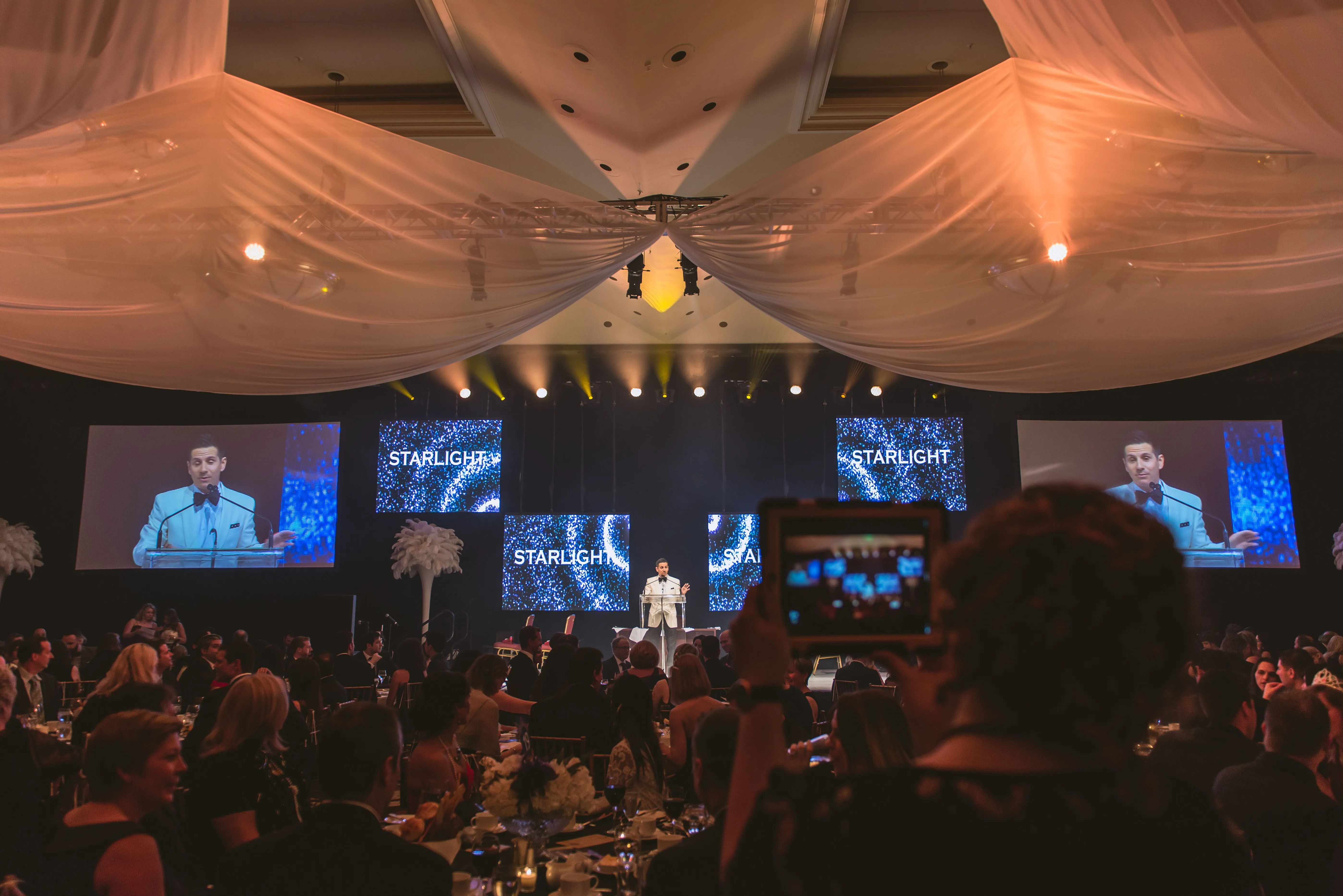 Man in white tuxedo speaking at podium on stage with 'STARLIGHT' displayed on multiple screens during formal event.