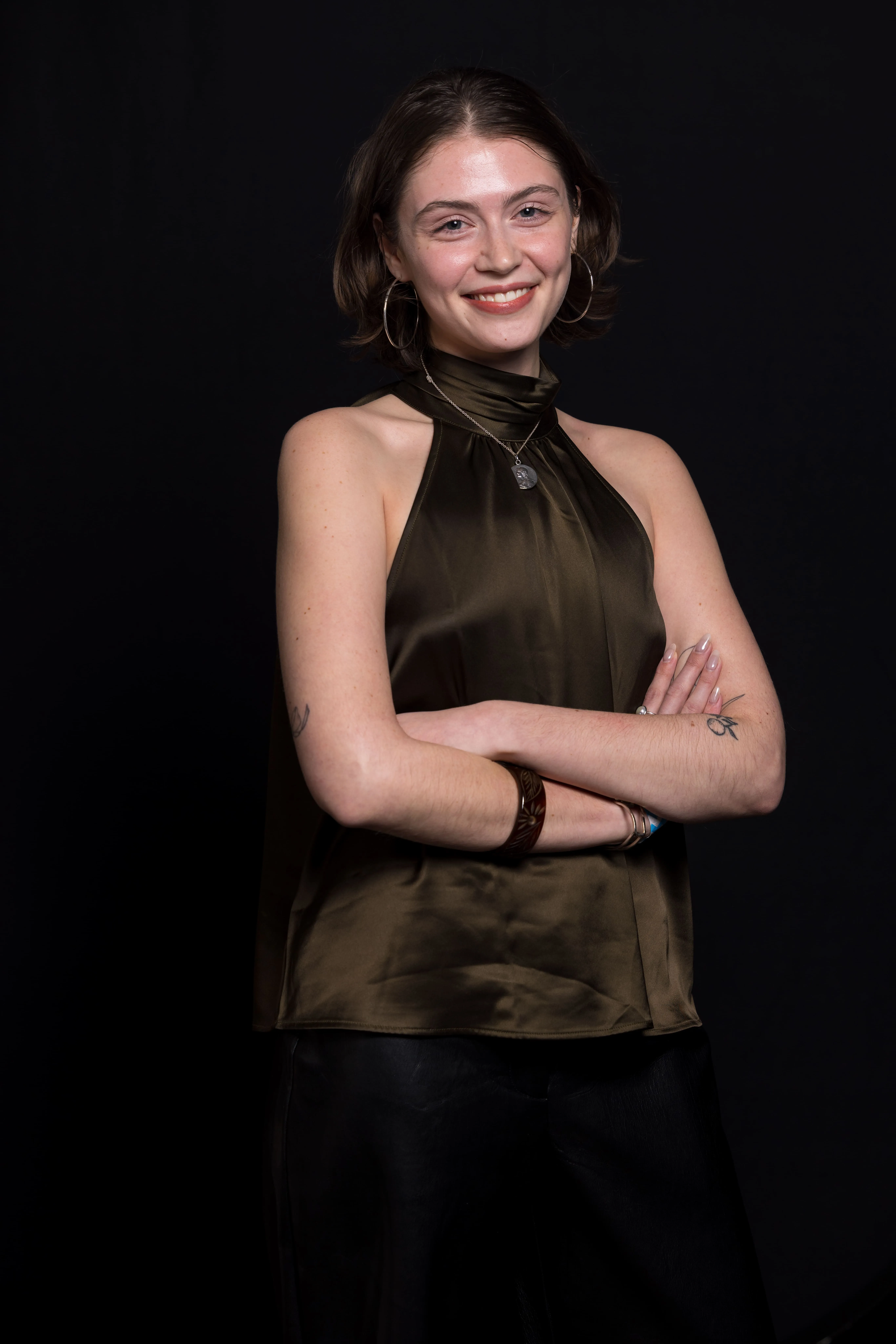 Young woman with short brown hair wearing a sleeveless olive green satin top, hoop earrings, and bracelets, smiling with arms crossed against a black background.