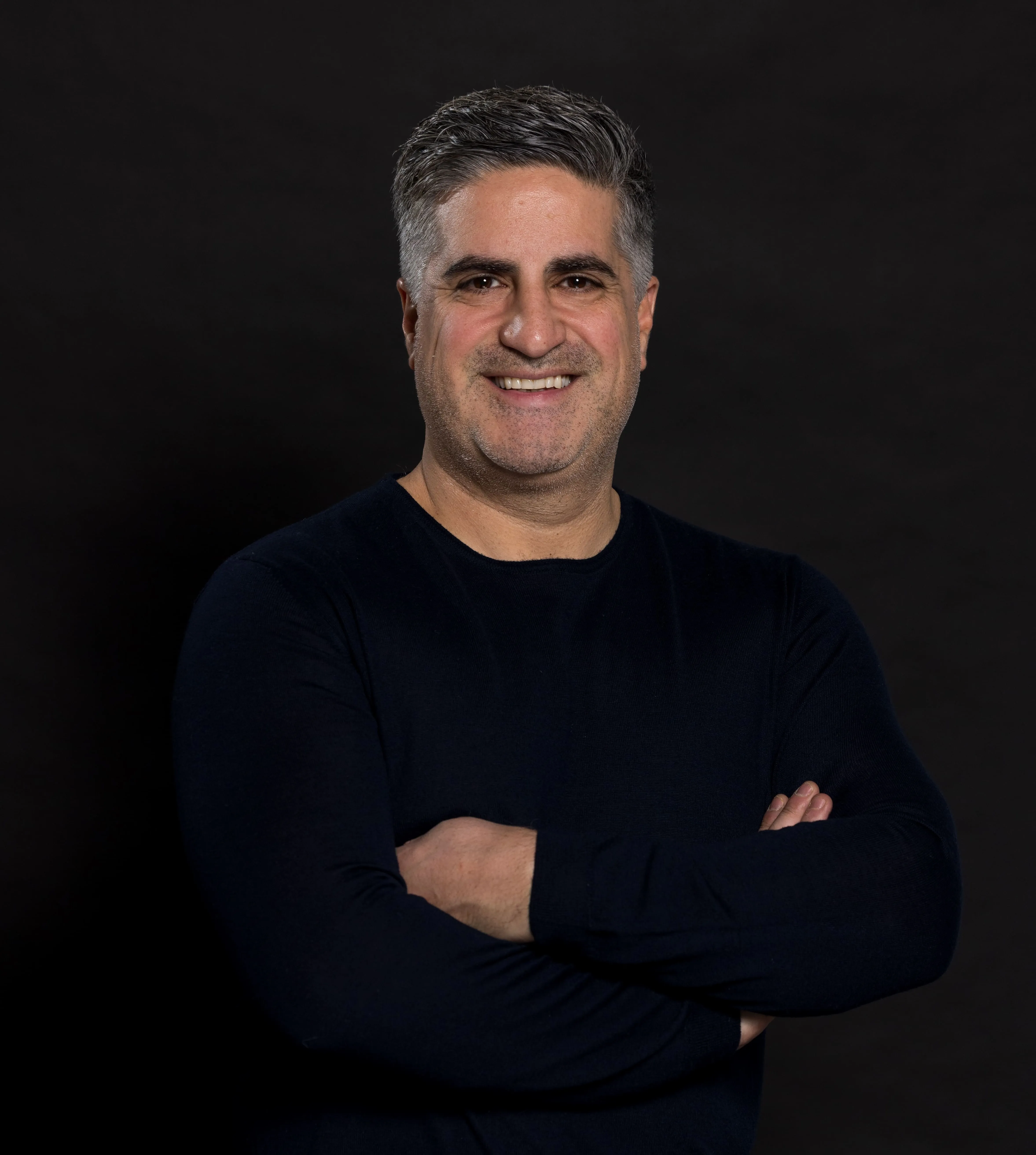 Smiling man with salt-and-pepper hair wearing a black long-sleeve shirt, arms crossed against a black background.