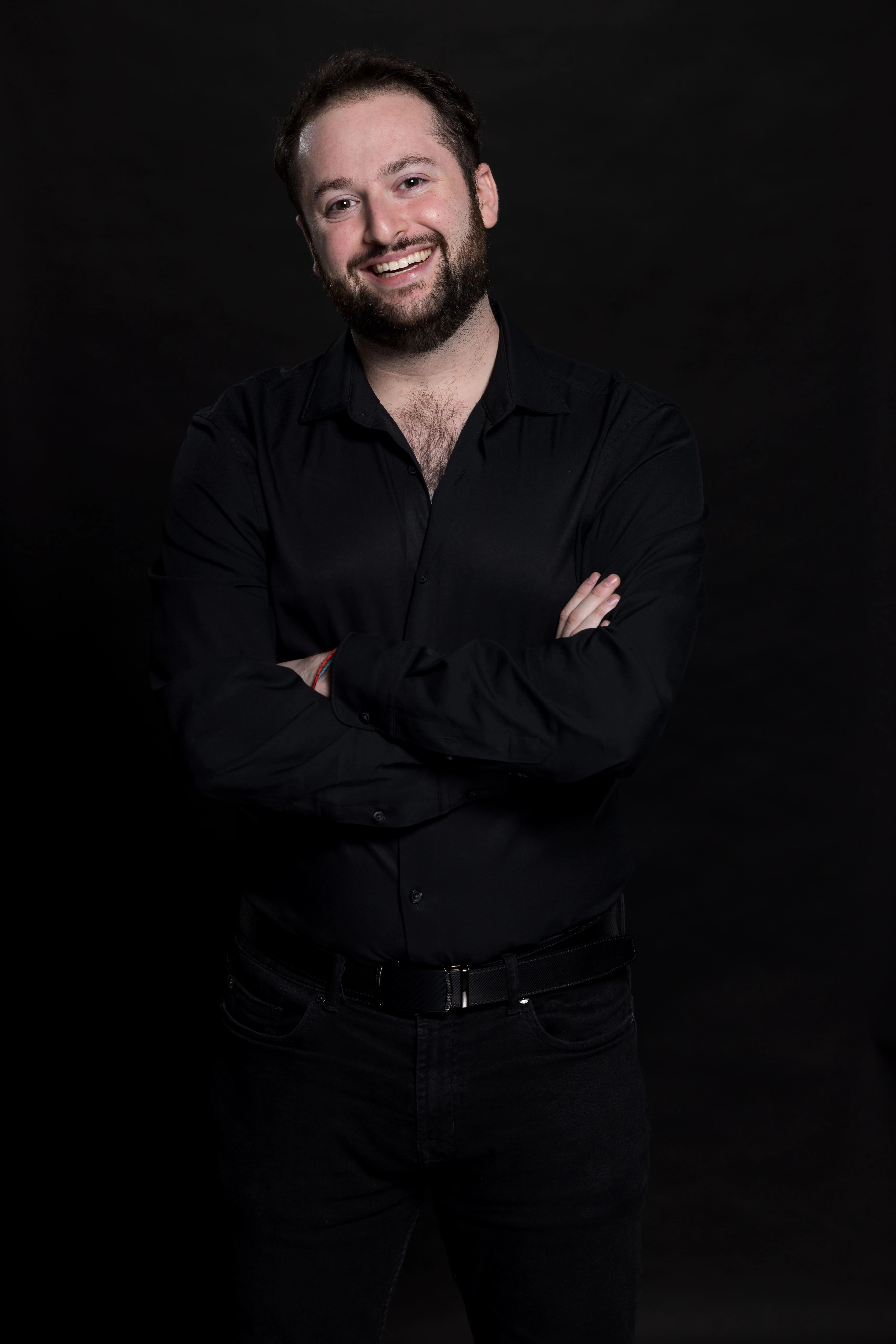 Smiling man with beard and short hair wearing a black shirt and black pants, standing with arms crossed against a black background.