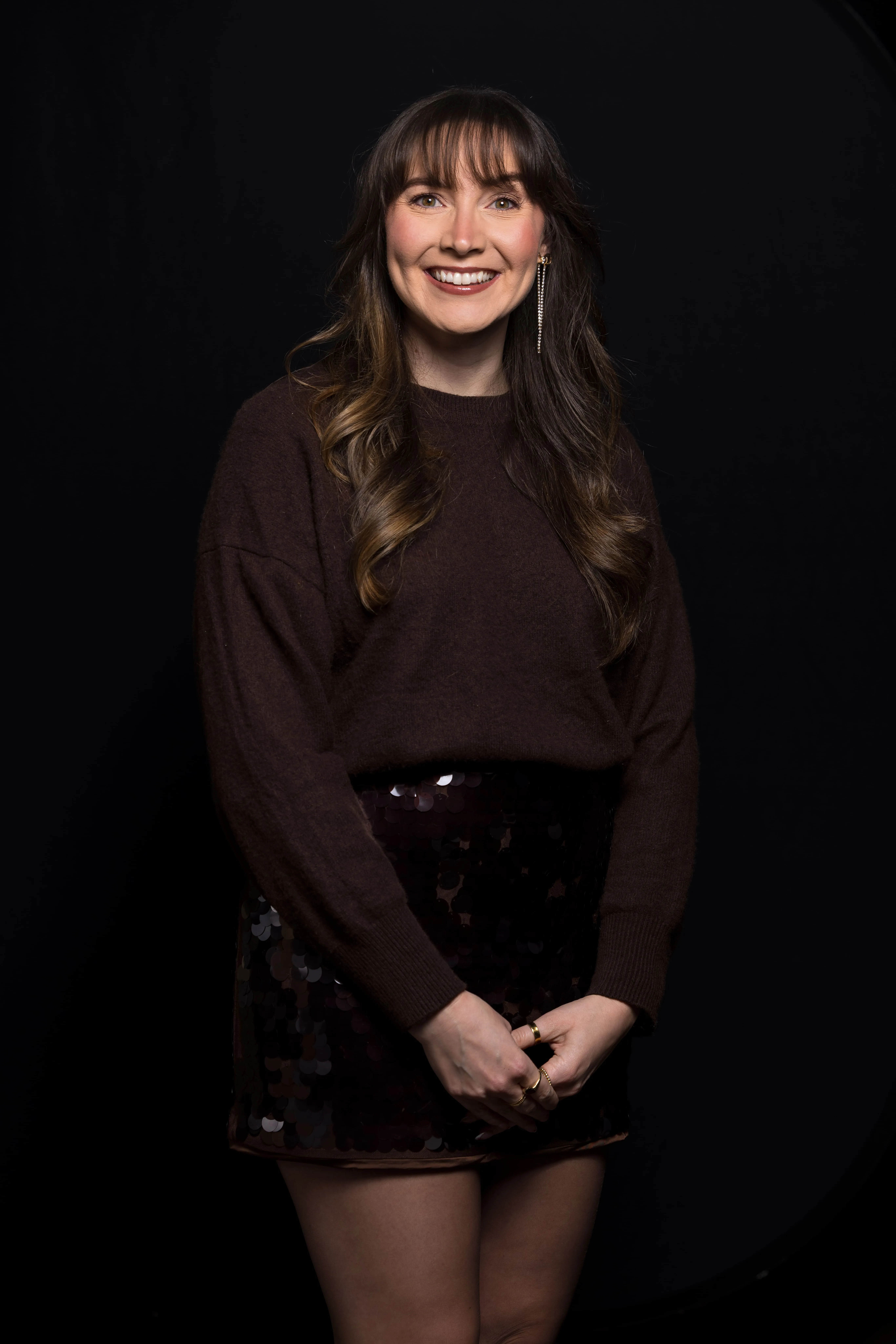 Smiling woman with long wavy hair wearing a dark brown sweater and a shiny black skirt, standing against a black background.