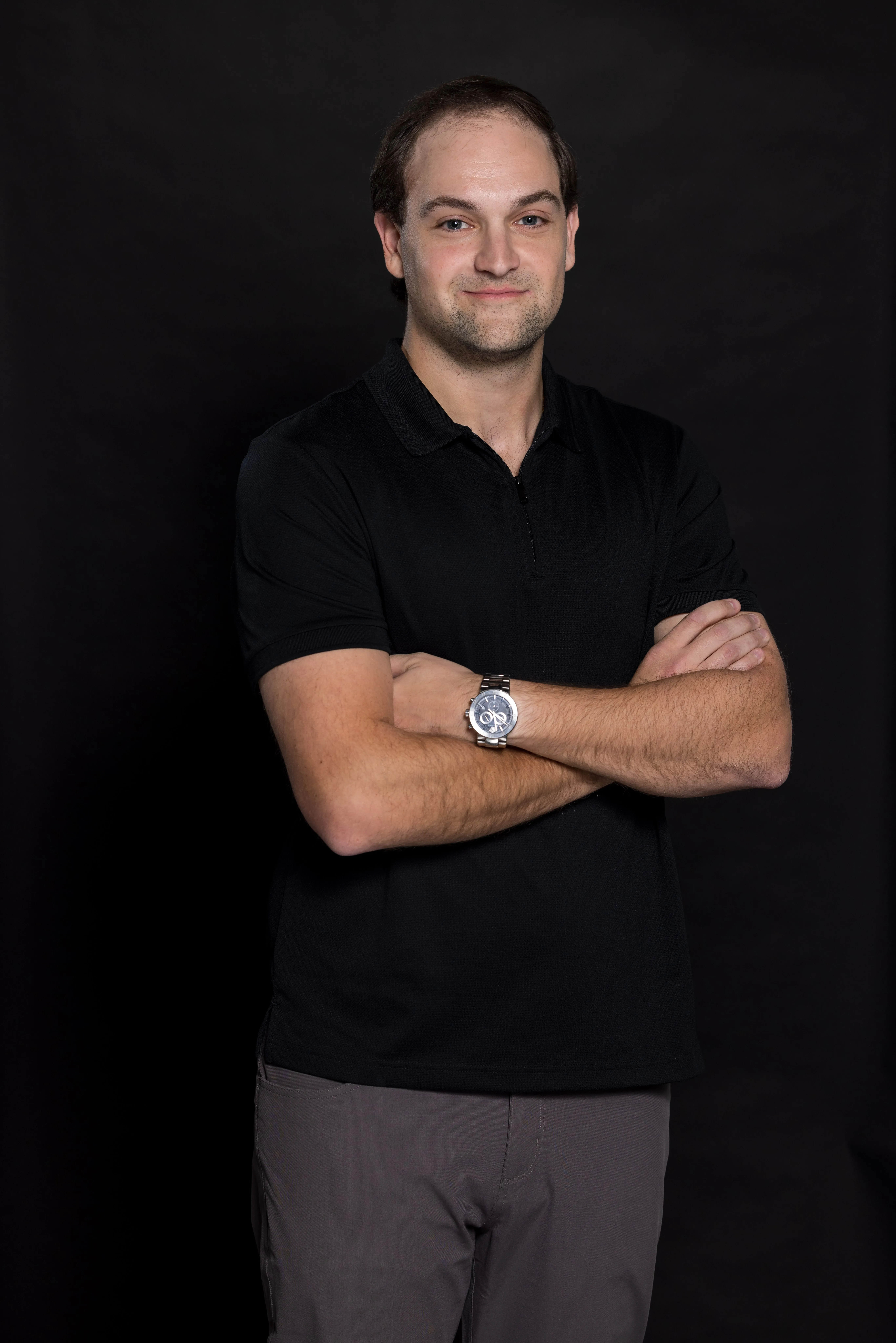 Man standing with arms crossed wearing a black polo shirt and gray pants against a black background.