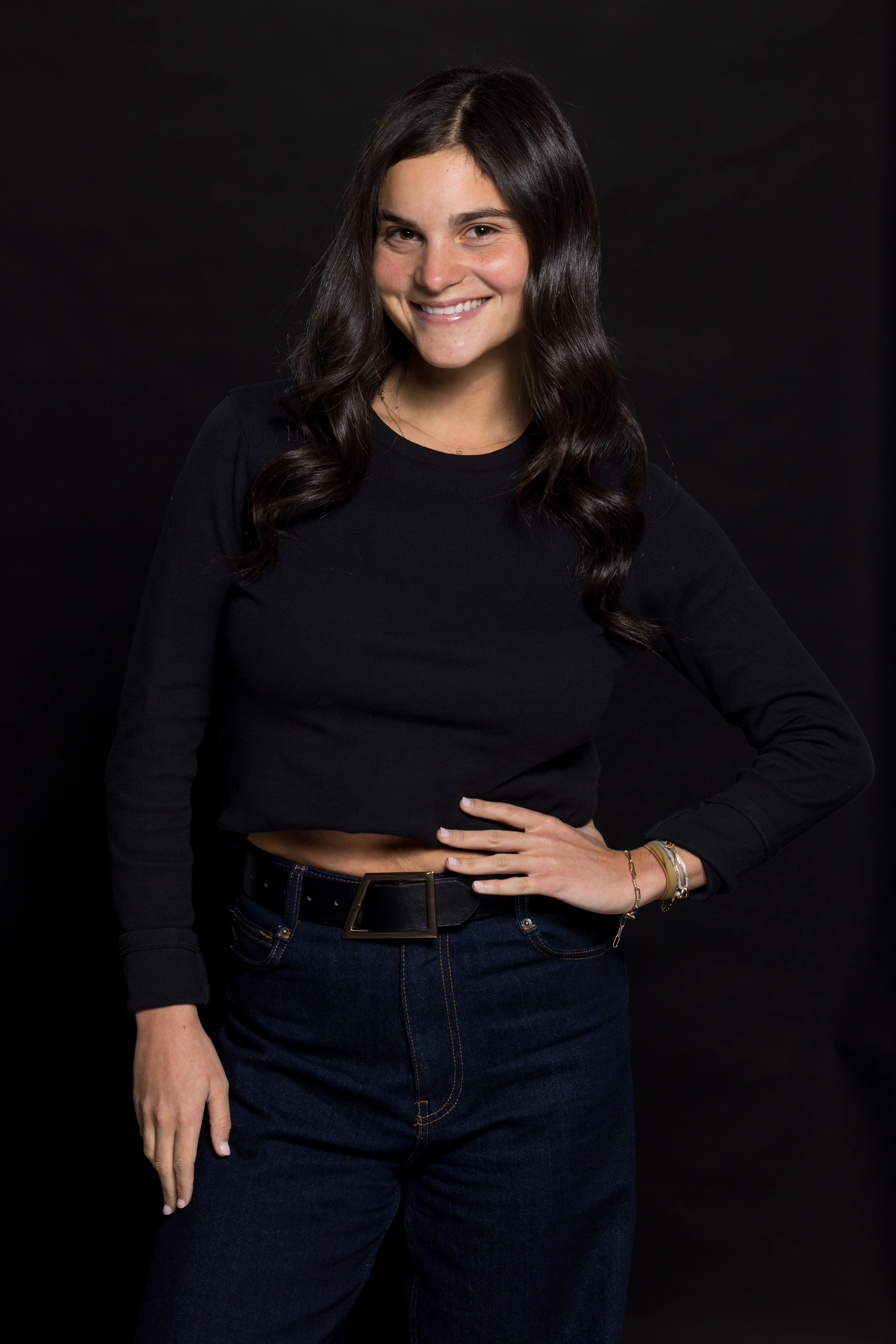 Smiling woman with long dark wavy hair wearing a black cropped sweater and dark jeans with a black belt against a black background.