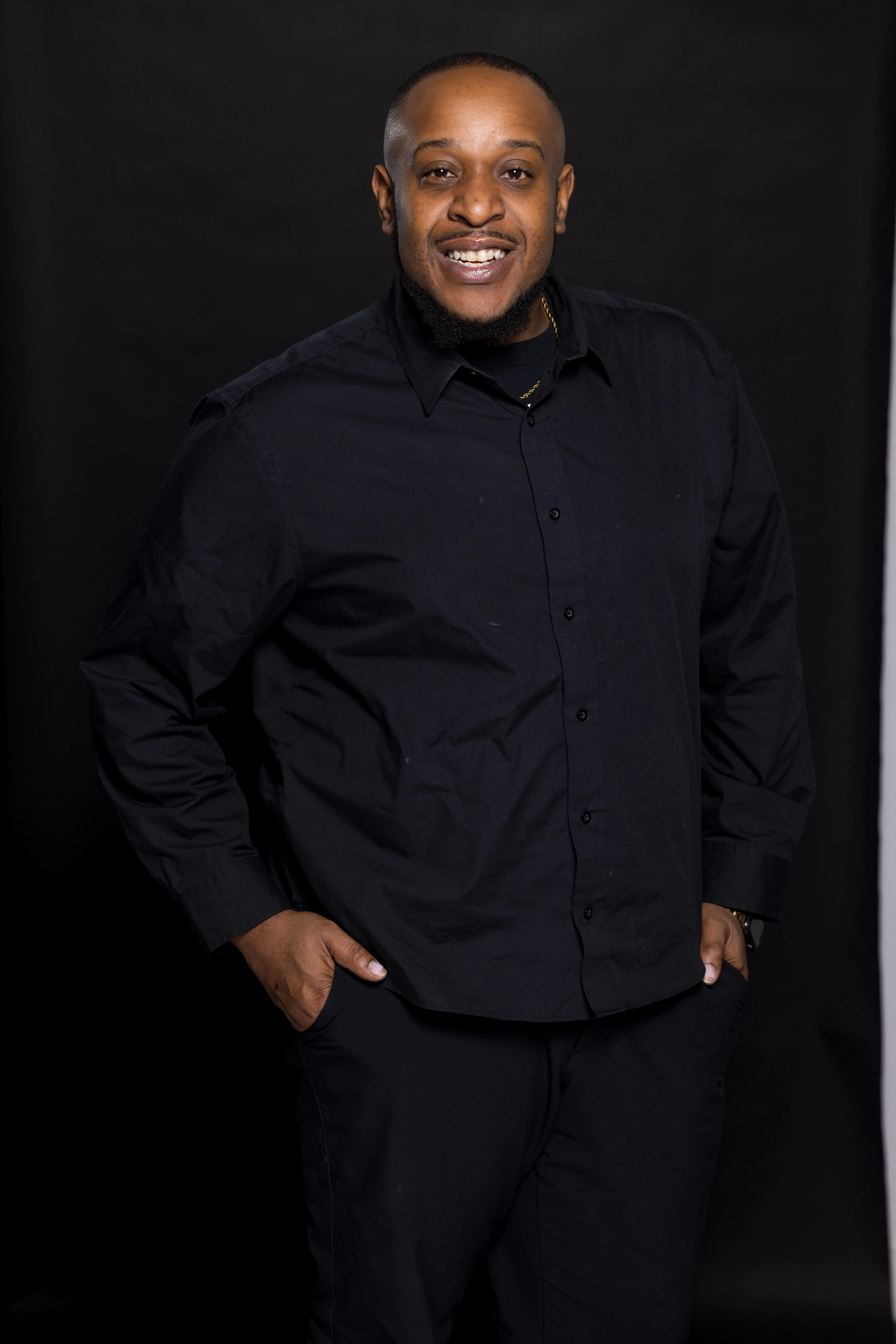 Smiling man with short hair and trimmed beard wearing a black button-up shirt and black pants, standing with hands in pockets against a black backdrop.