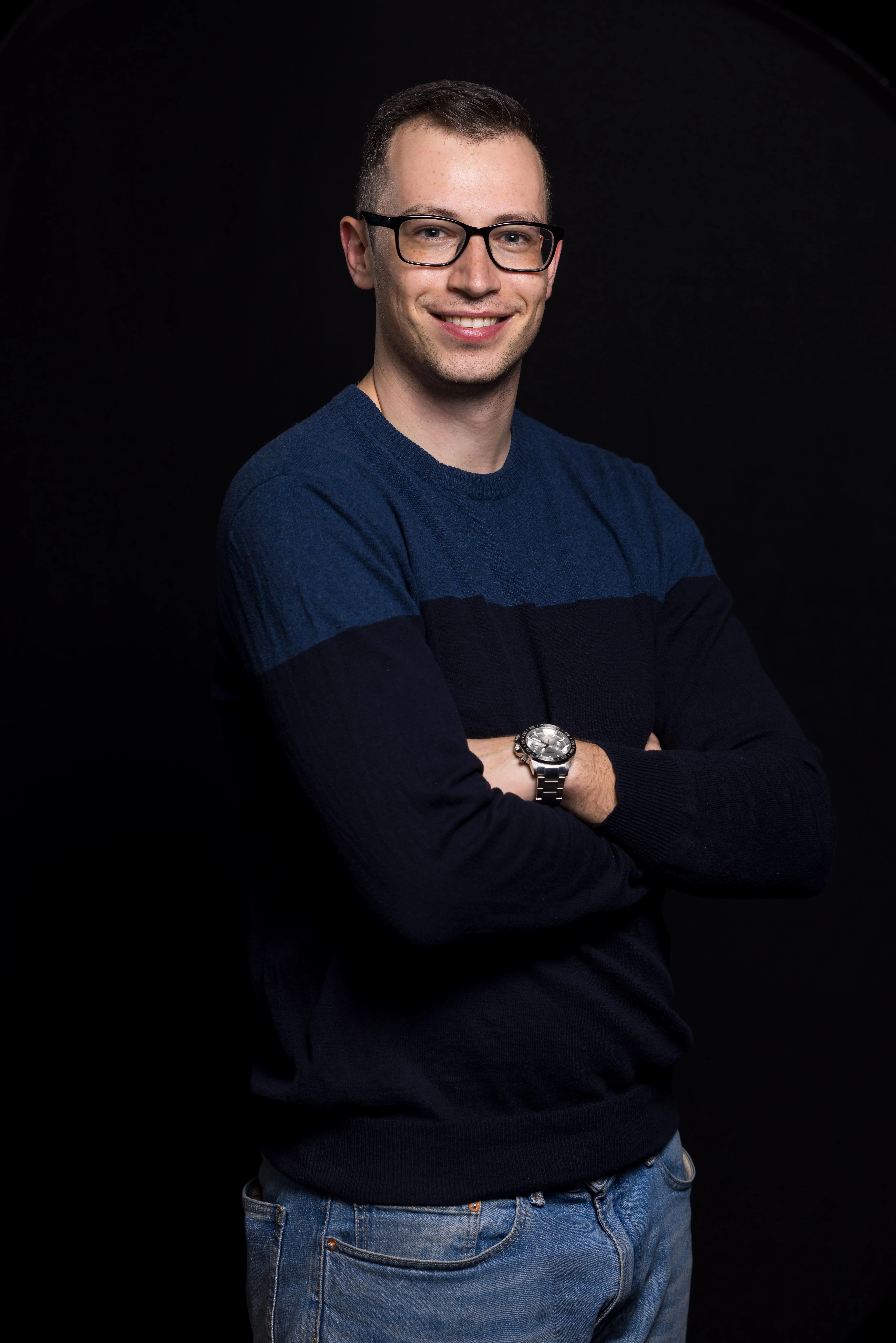 Smiling man with glasses wearing a blue and black sweater and jeans, arms crossed against black background.