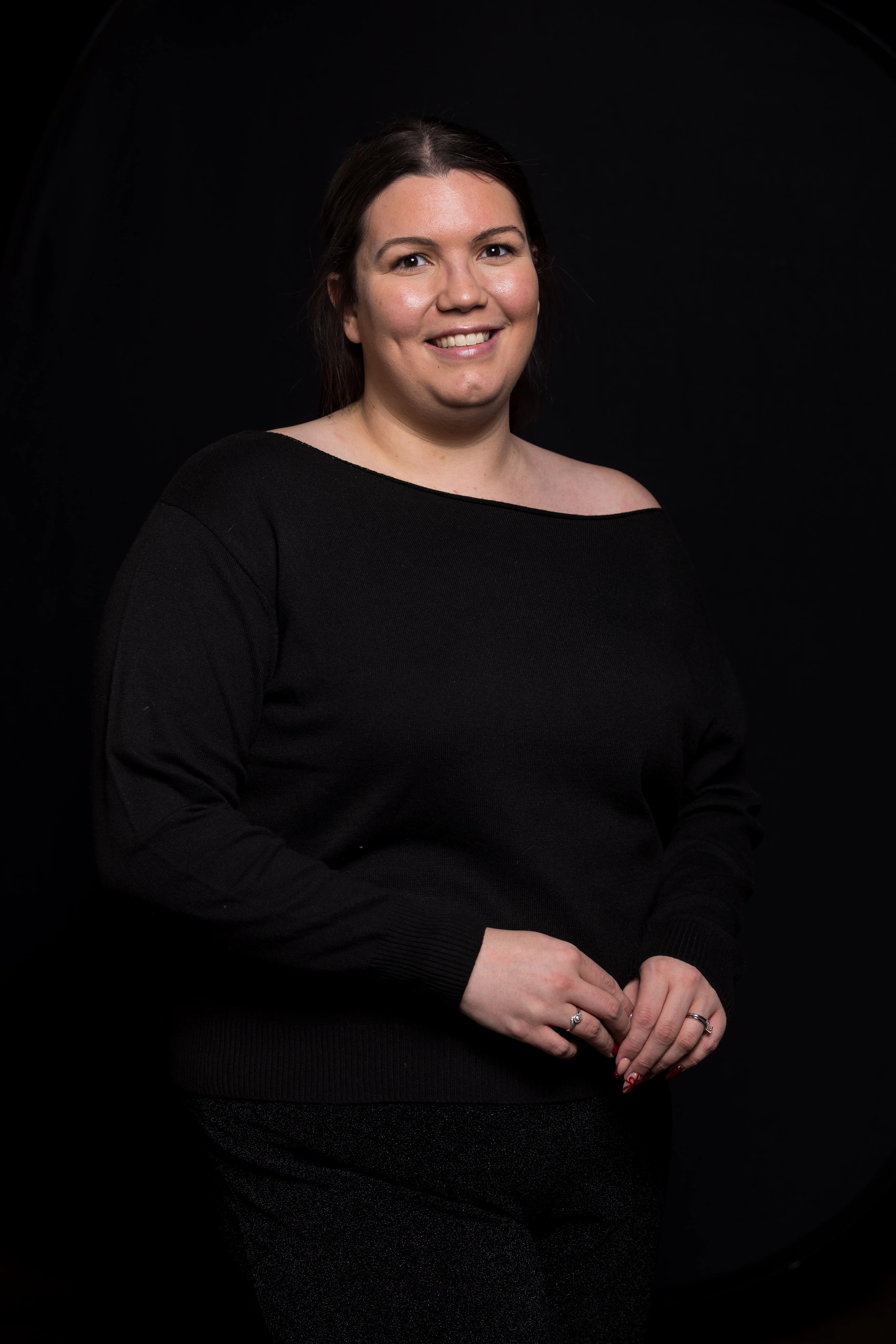 Smiling woman with dark hair in a black off-shoulder sweater against a black background.