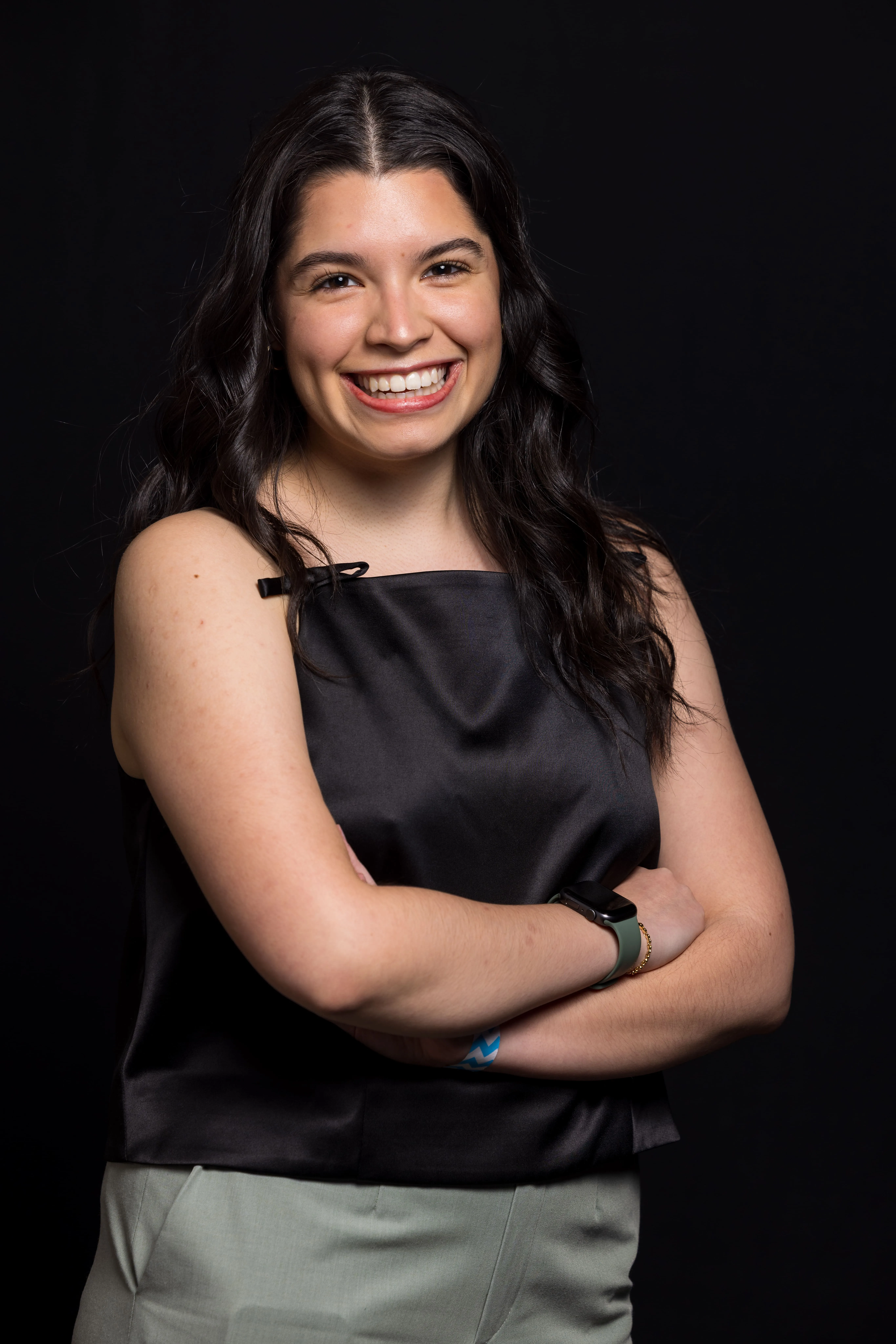 Smiling woman with long dark hair wearing a black sleeveless top and light gray pants, standing with crossed arms against a black background.