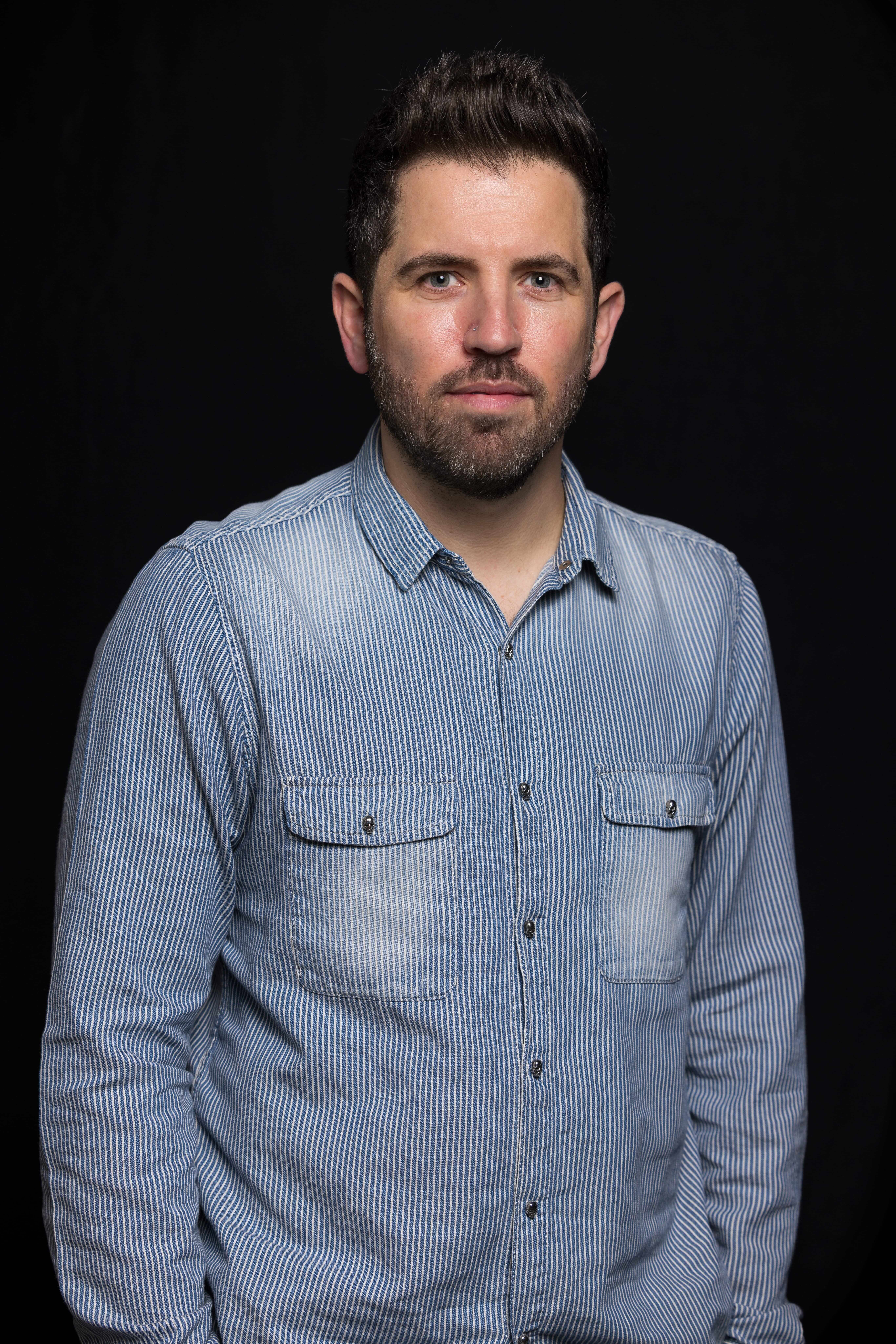 Man with short dark hair and beard wearing a blue and white striped button-up shirt against a black background.