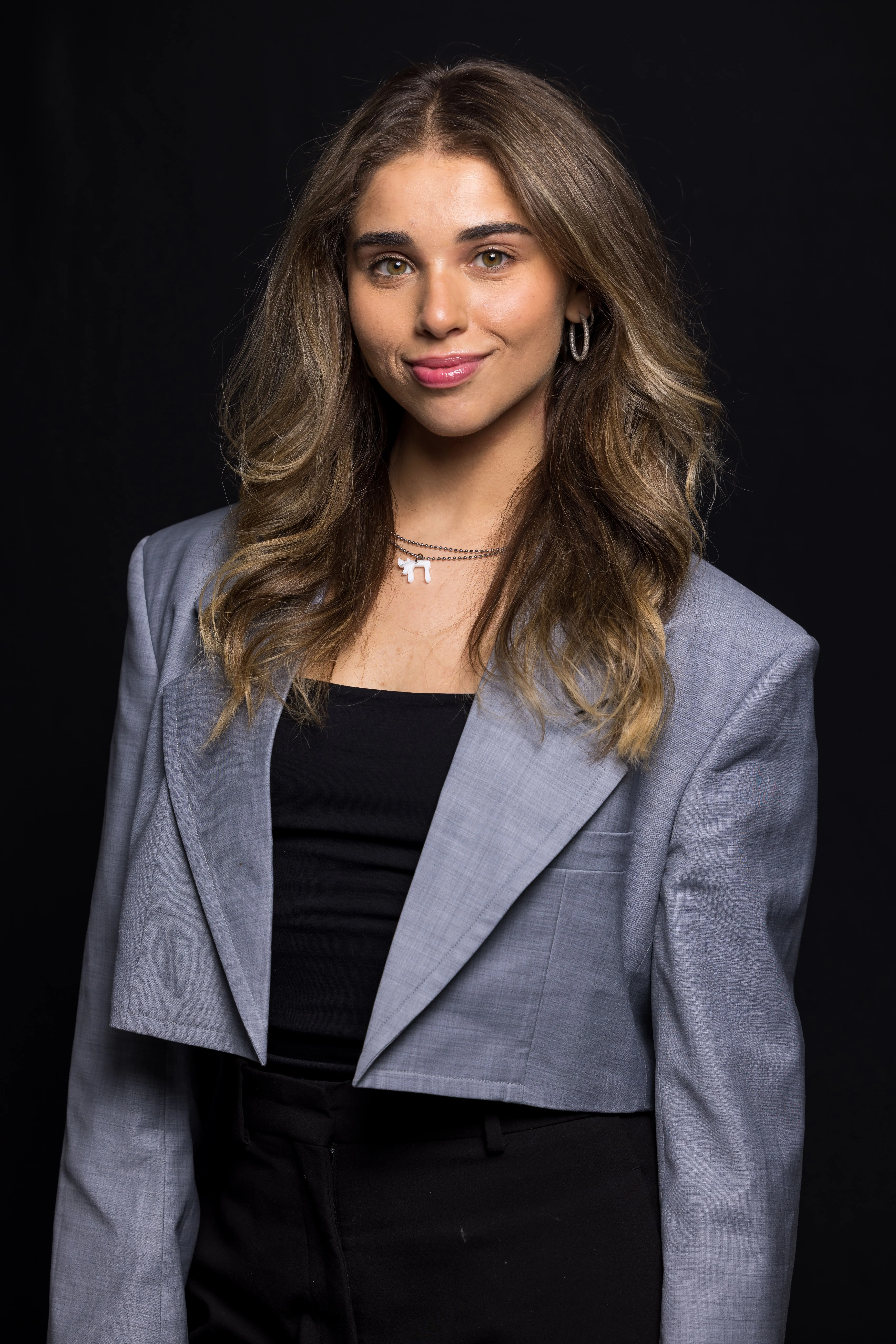 Young woman with long wavy hair wearing a gray cropped blazer, black top, and silver hoop earrings against a black background.