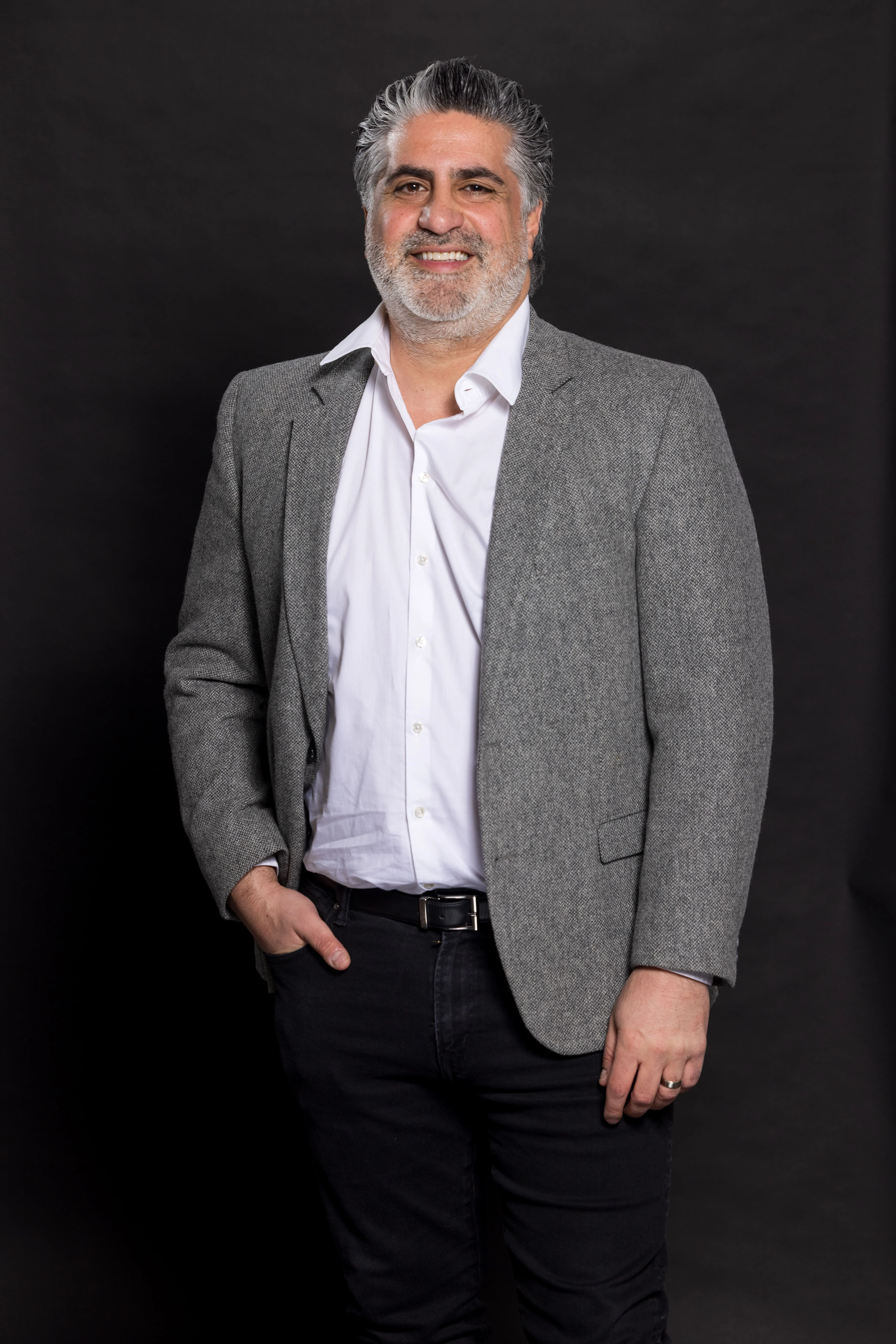 Smiling middle-aged man with gray hair and beard wearing a gray blazer over a white shirt and black pants with one hand in pocket against a dark background.