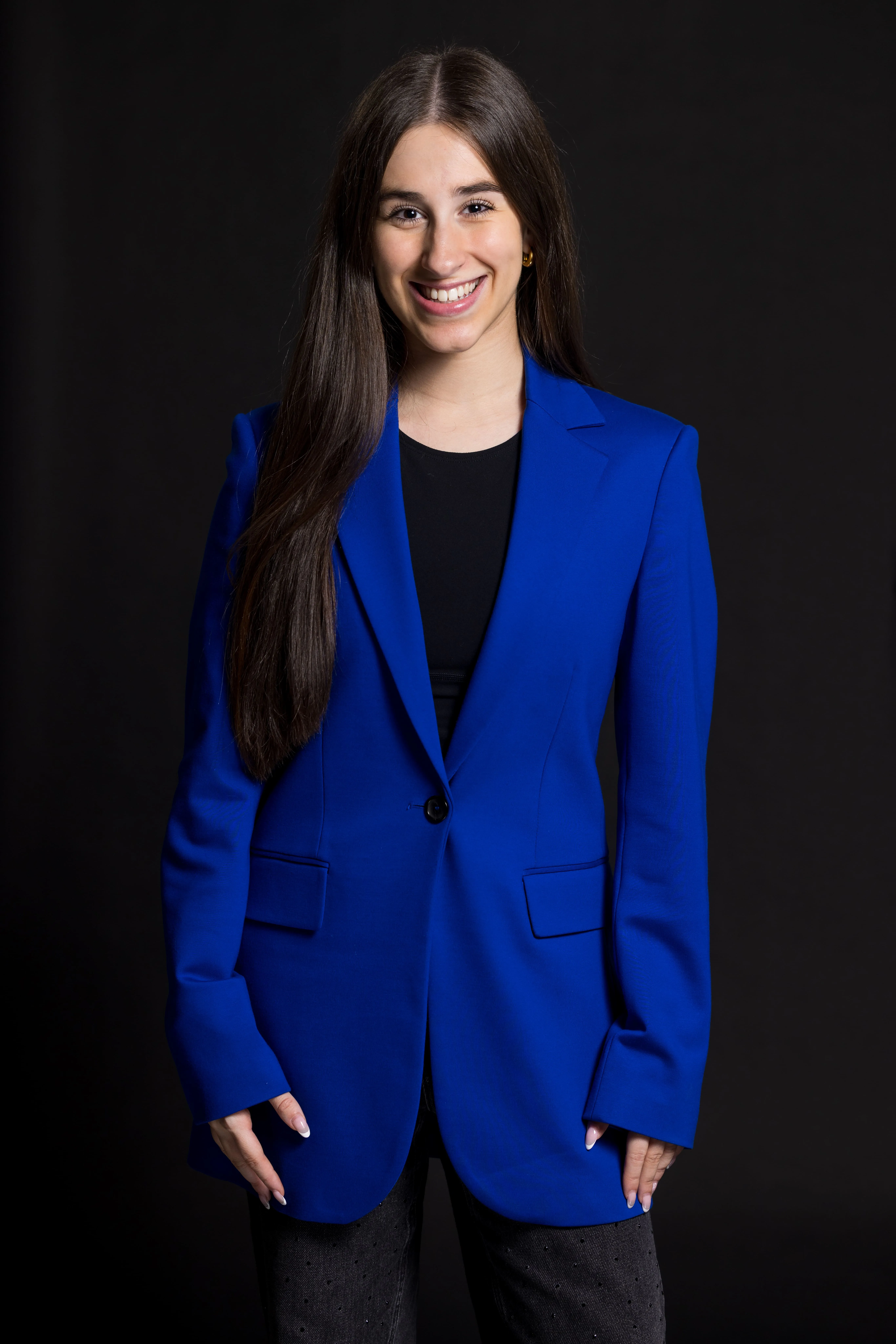 Smiling woman with long straight brown hair wearing a bright blue blazer over a black top against a black background.