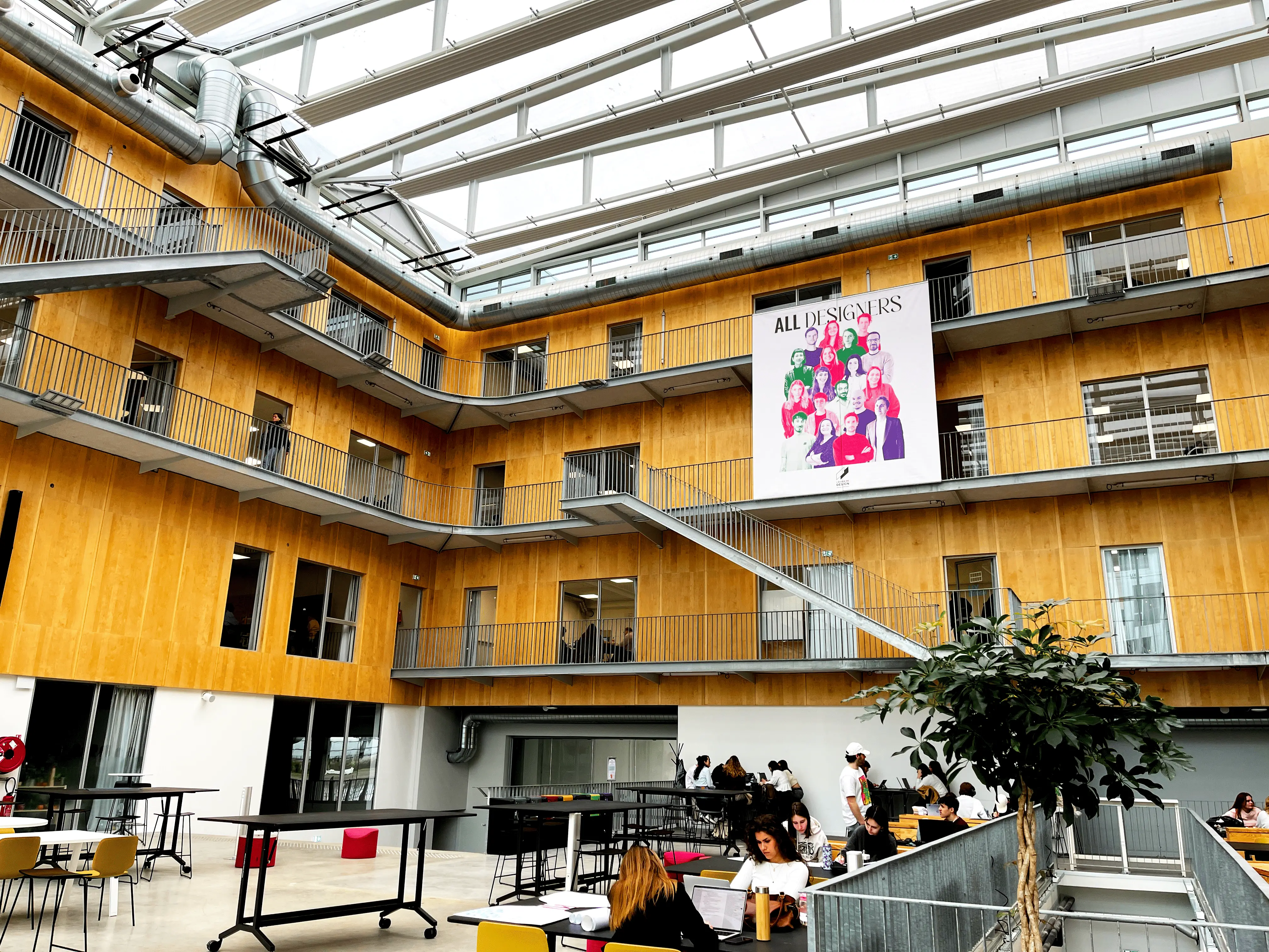 Intérieur lumineux de l'école de design Nantes Atlantique avec plusieurs étages en bois clair, un grand escalier métallique et des étudiants travaillant à des tables dans un espace ouvert.