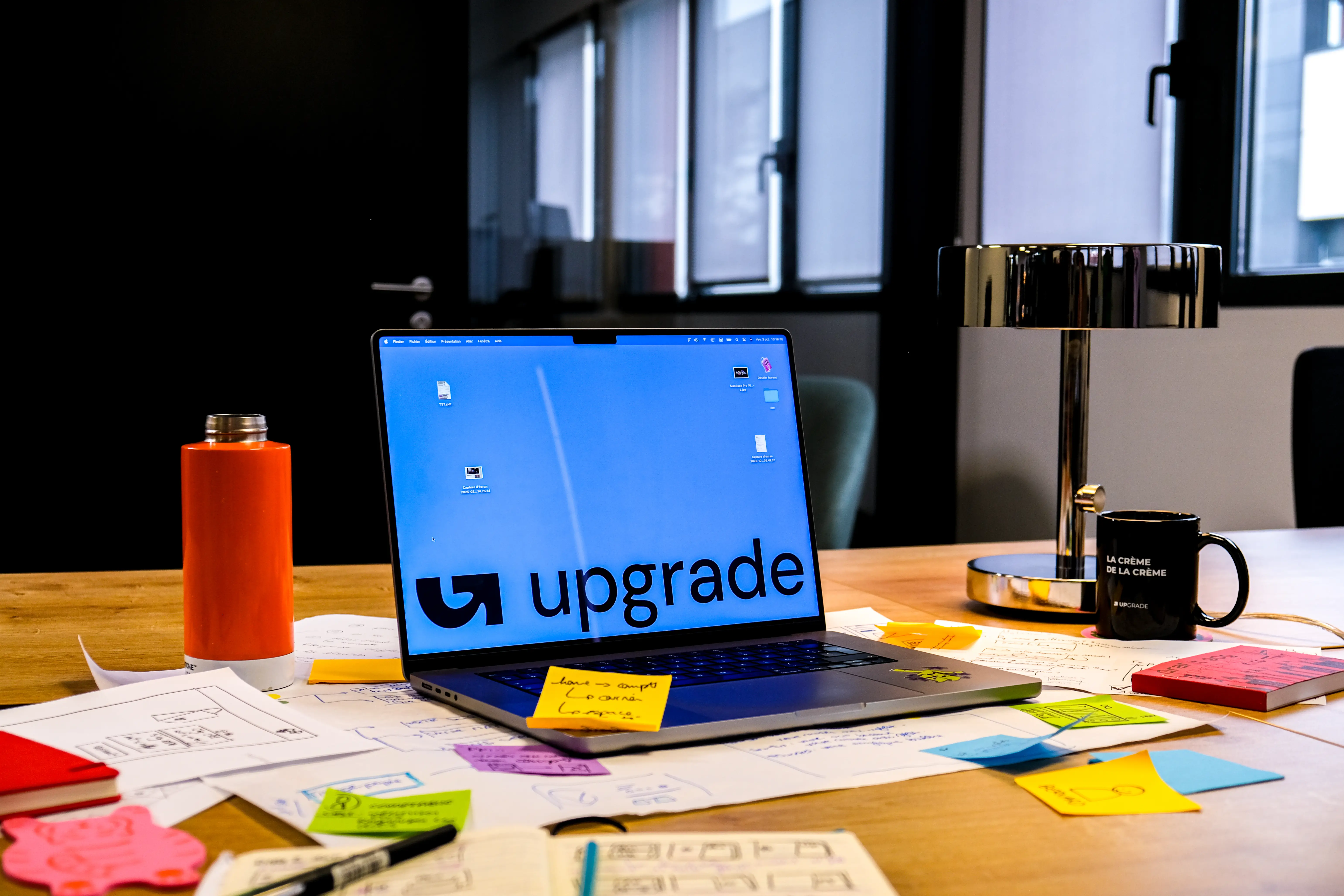 Vue d'un poste de travail chez l'agence digitale Upgrade, avec des outils de conception (croquis, Post-it) et le logo Upgrade à l'écran, reflétant l'expertise de notre agence design UX UI Nantes Lyon Paris.