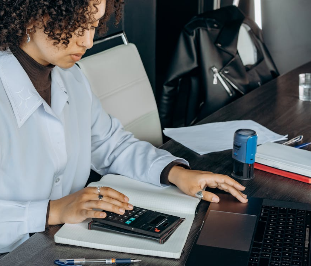 Person using a calculator and laptop at a desk with papers and a stamp nearby.