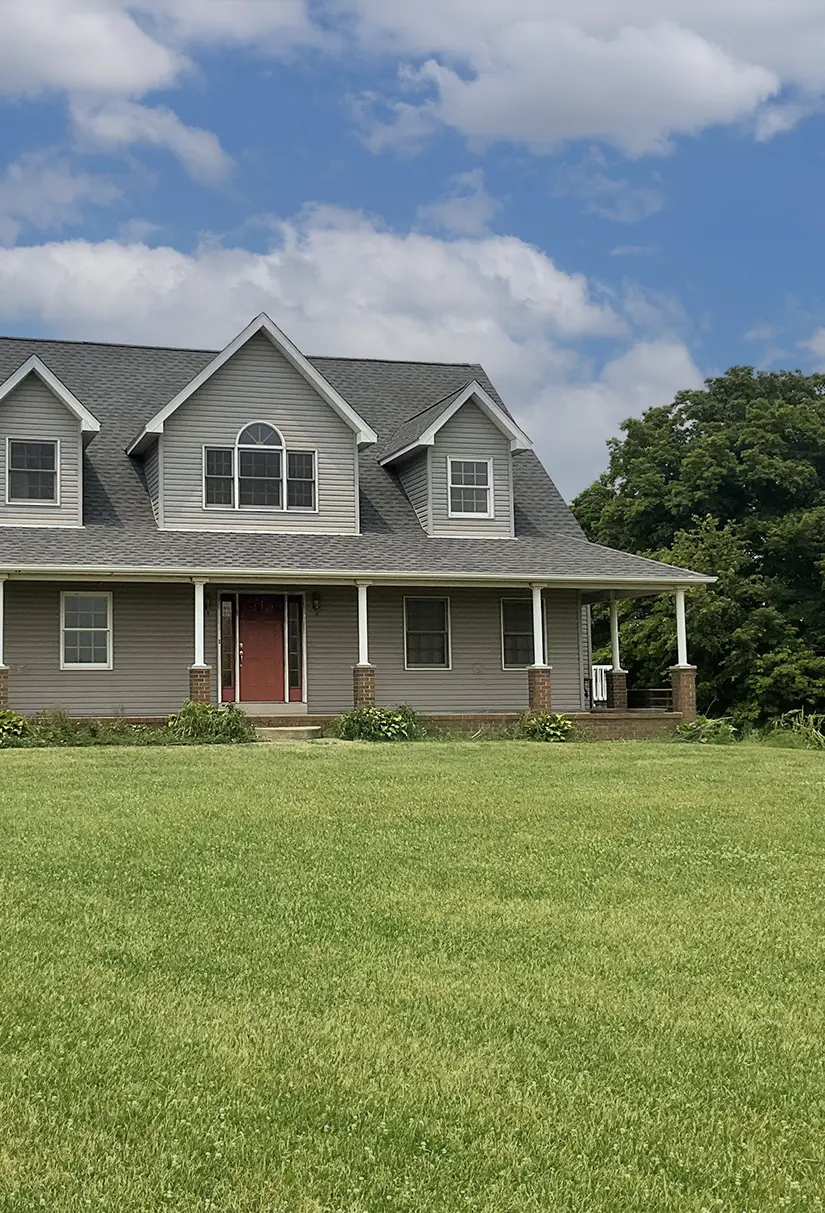 Gray two-story house with a red front door, multiple windows, a porch with white columns, surrounded by green lawn and trees under a partly cloudy sky.
