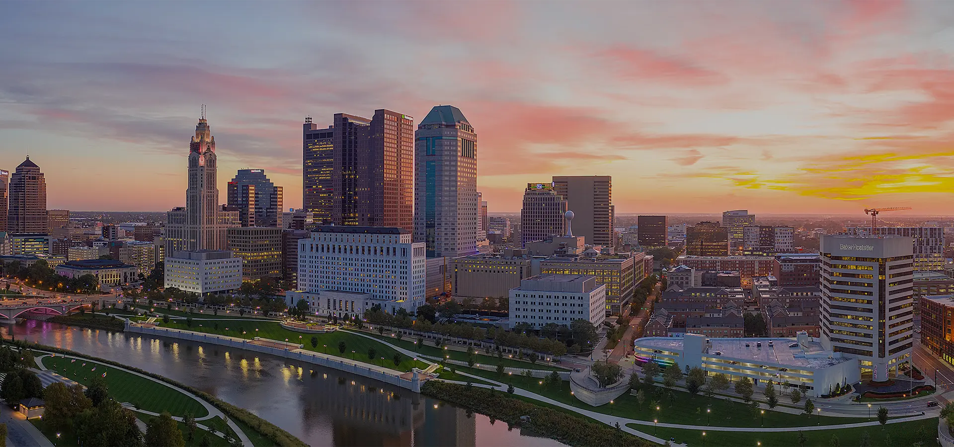 Panoramic view of Columbus, Ohio skyline at sunset with river and illuminated buildings.