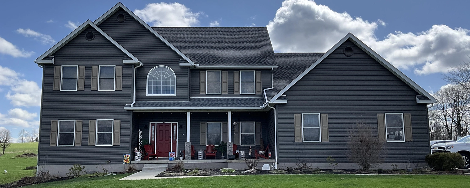 Two-story house with dark gray siding, brown shutters, a red front door, and a large front porch with chairs.