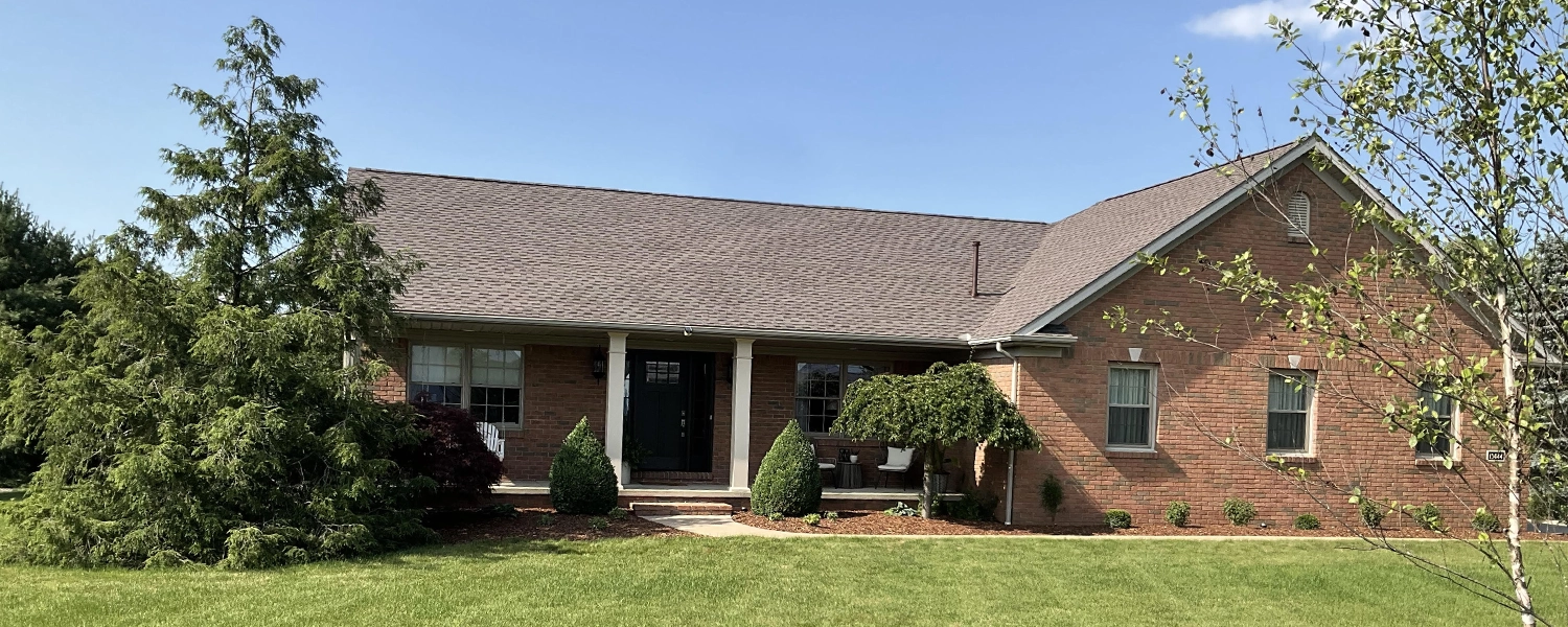 Brick house with a well-maintained shingle roof, front porch, shrubs, and green lawn under a clear blue sky.