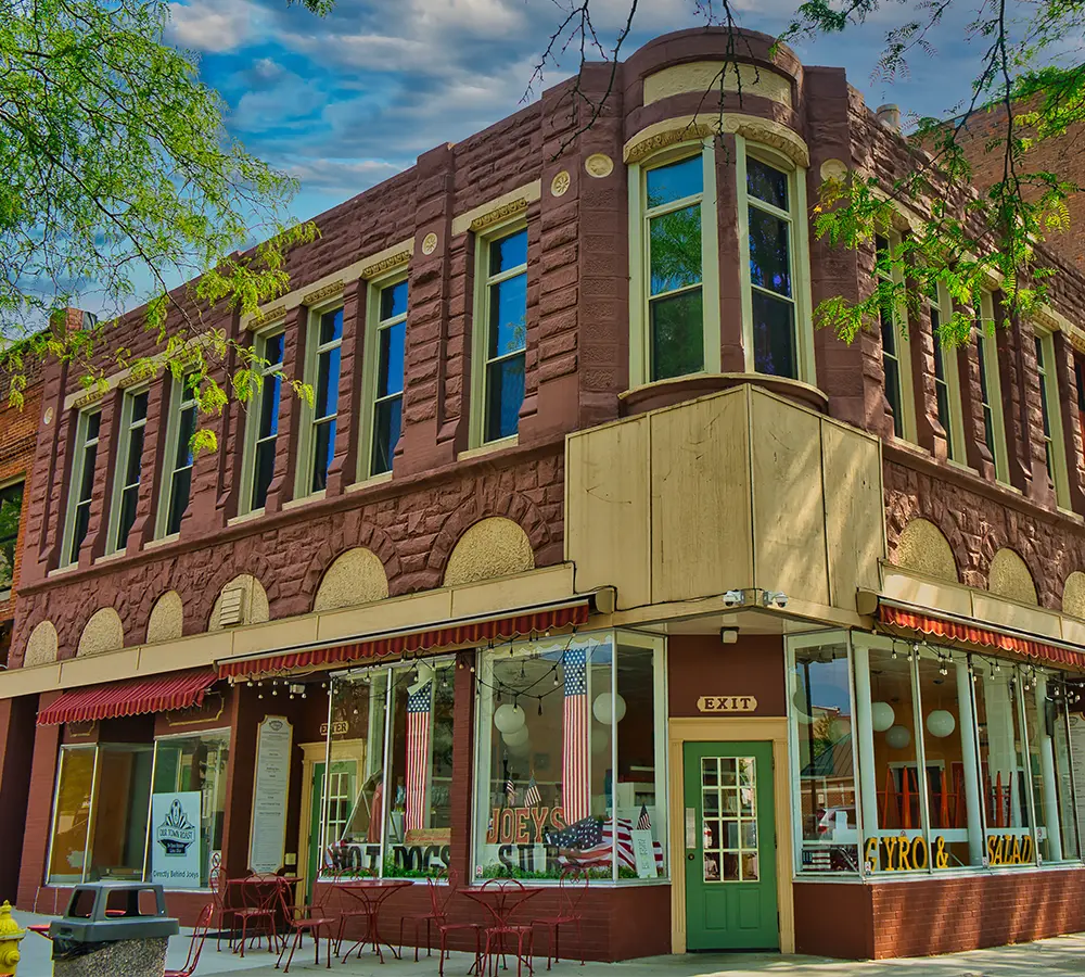 Historic two-story brick building with large storefront windows displaying American flags and signs for hot dogs, gyro, and salad, with outdoor tables on the sidewalk.