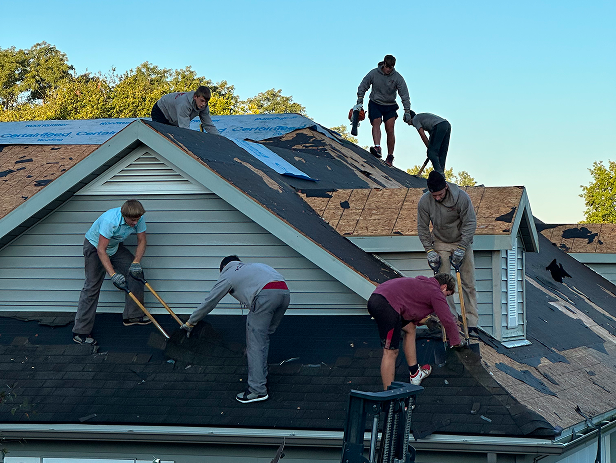 Six workers repairing and installing shingles on a sloped house roof under clear sky.