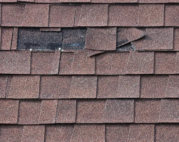 Close-up of a brown shingle roof with damaged and lifted shingles revealing underlying black layer.