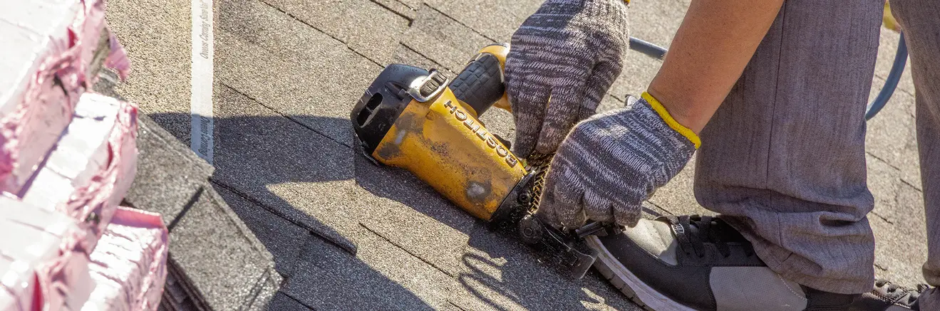 Person wearing gloves using a nail gun to install asphalt shingles on a roof.