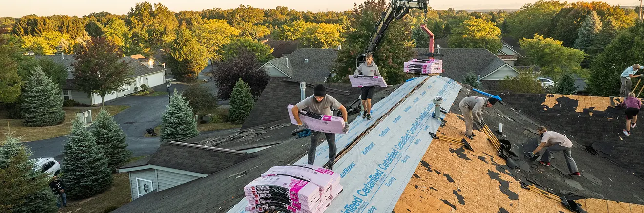 Roofers installing new shingles on a large sloped roof with trees and houses in the background.