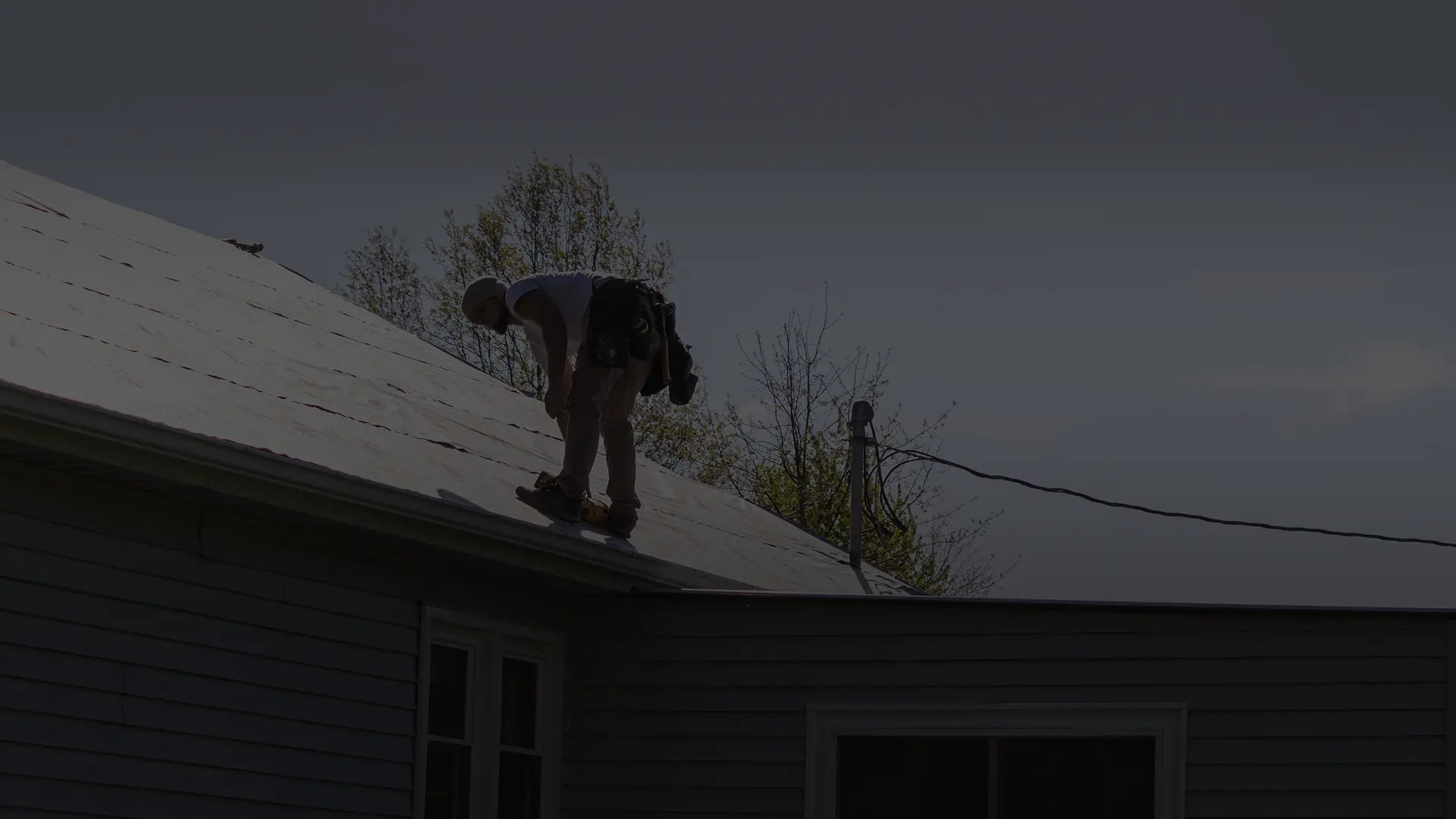 Worker wearing helmet using nail gun to install roofing material on a house roof.