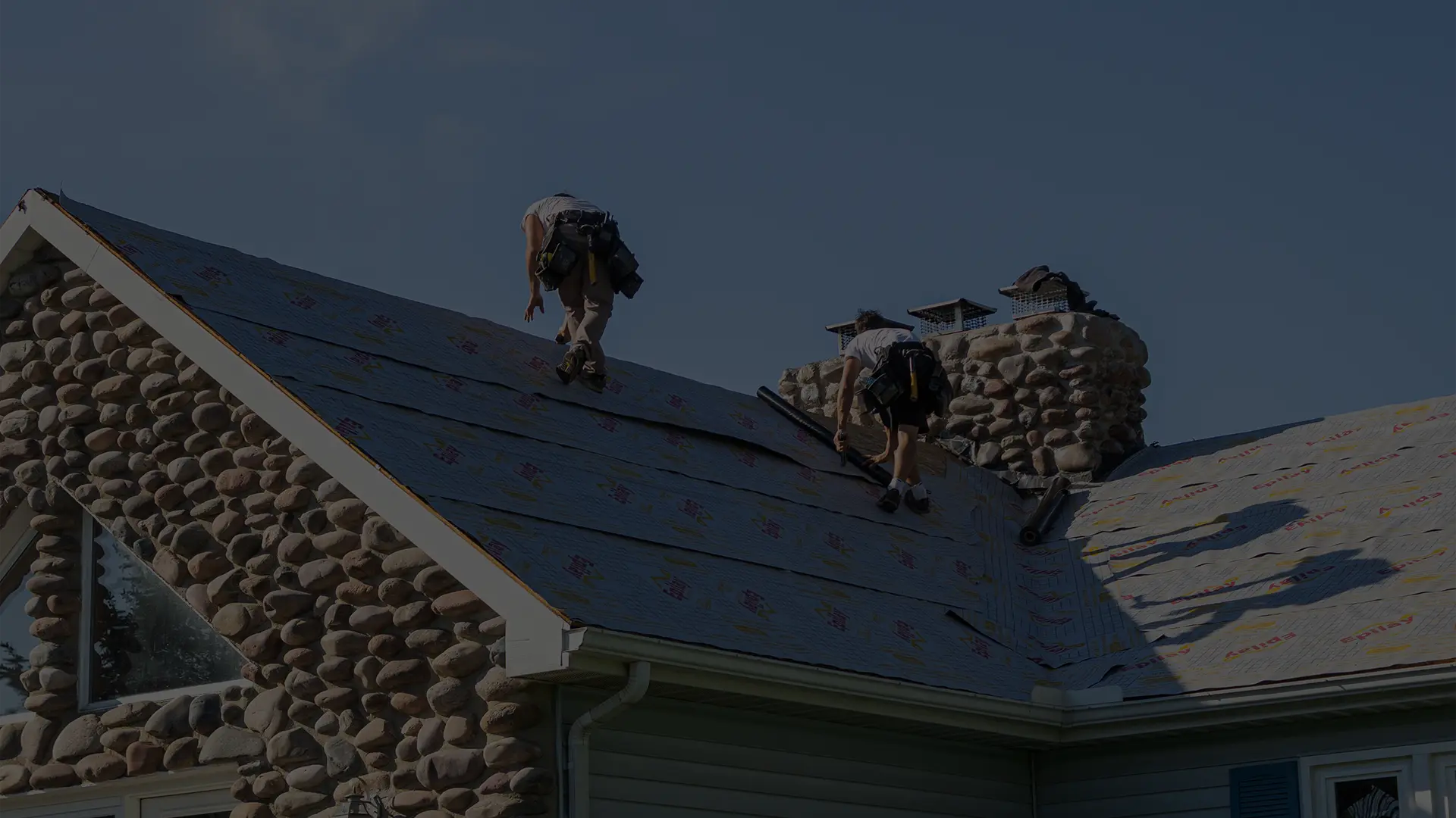 Worker wearing helmet using nail gun to install roofing material on a house roof.