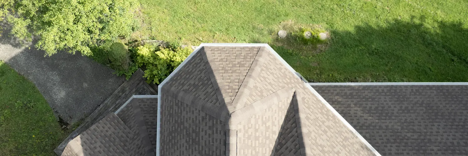 Aerial view of a rooftop with asphalt shingles surrounded by green grass and trees.