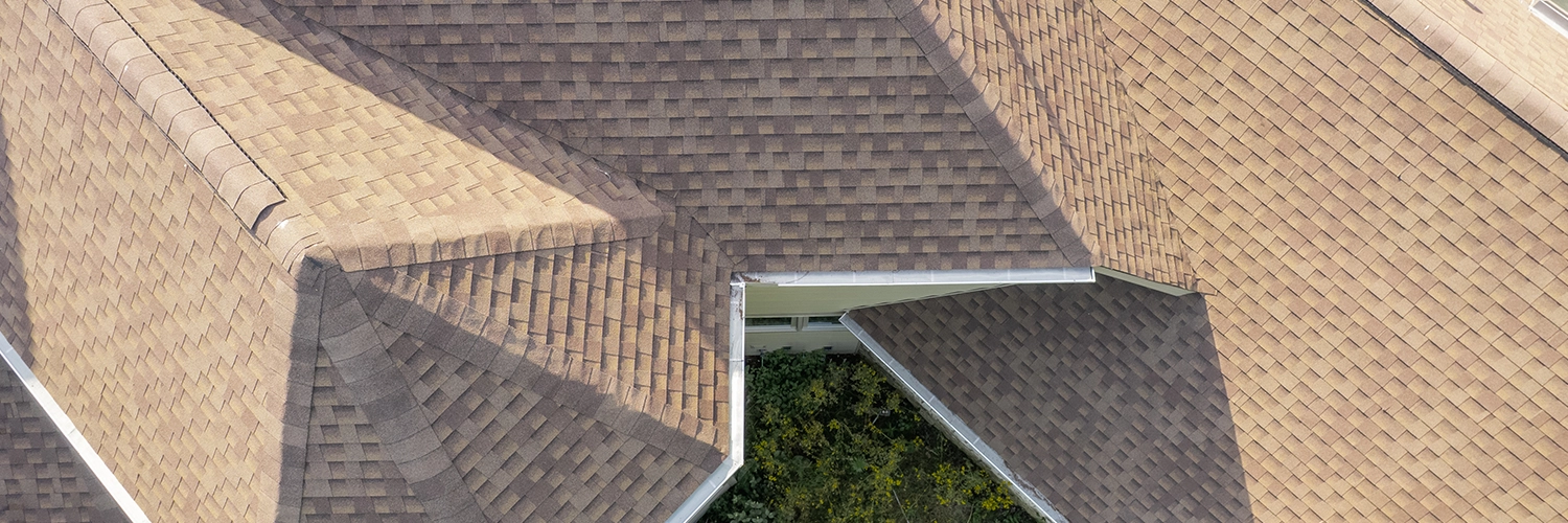 Aerial view of multiple brown asphalt shingle roofs with valleys and ridges surrounded by green foliage.