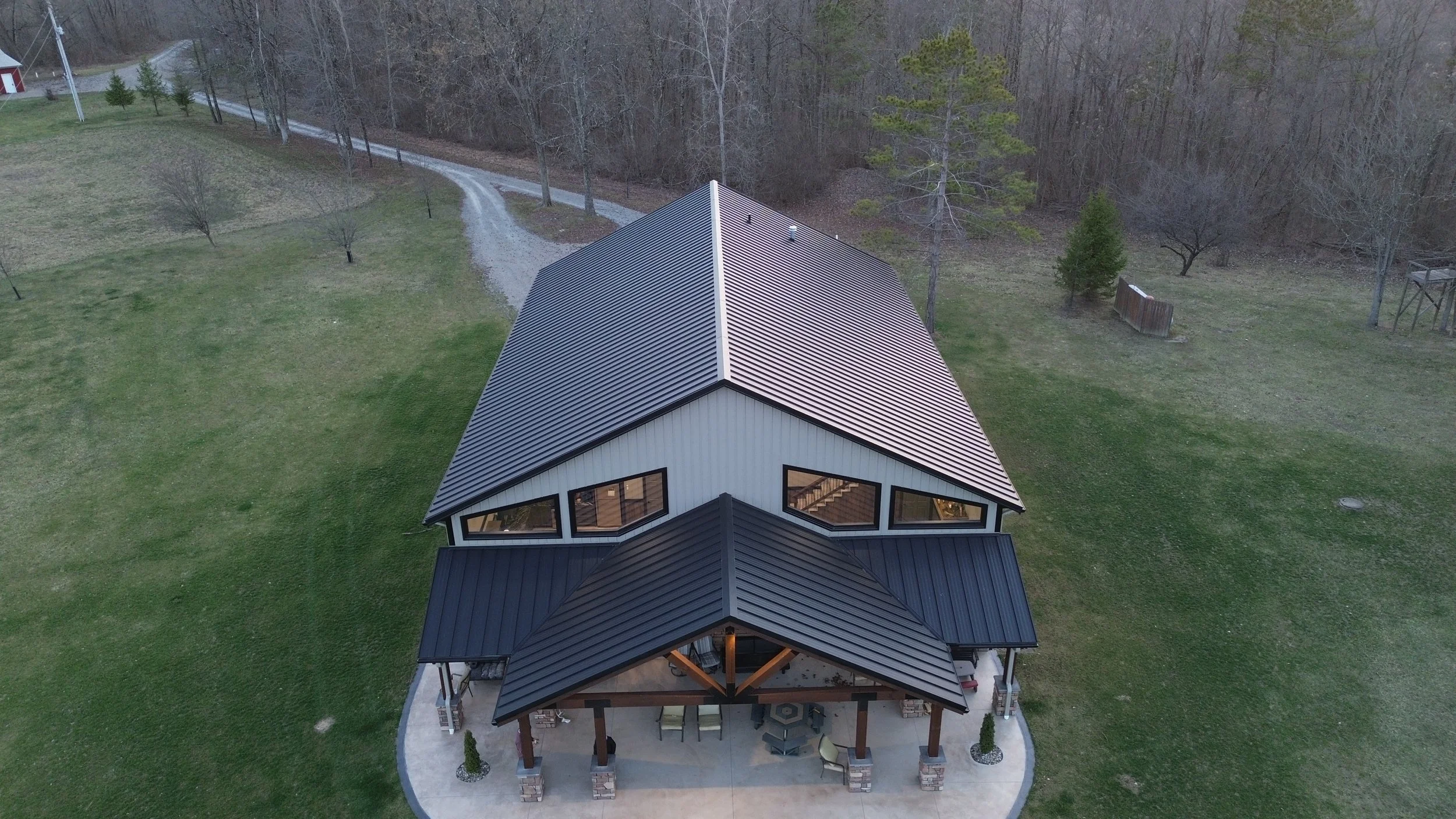 A modern house with a dark metal standing seam roof, large triangular windows, and a covered front porch with furniture, surrounded by lawn and trees.