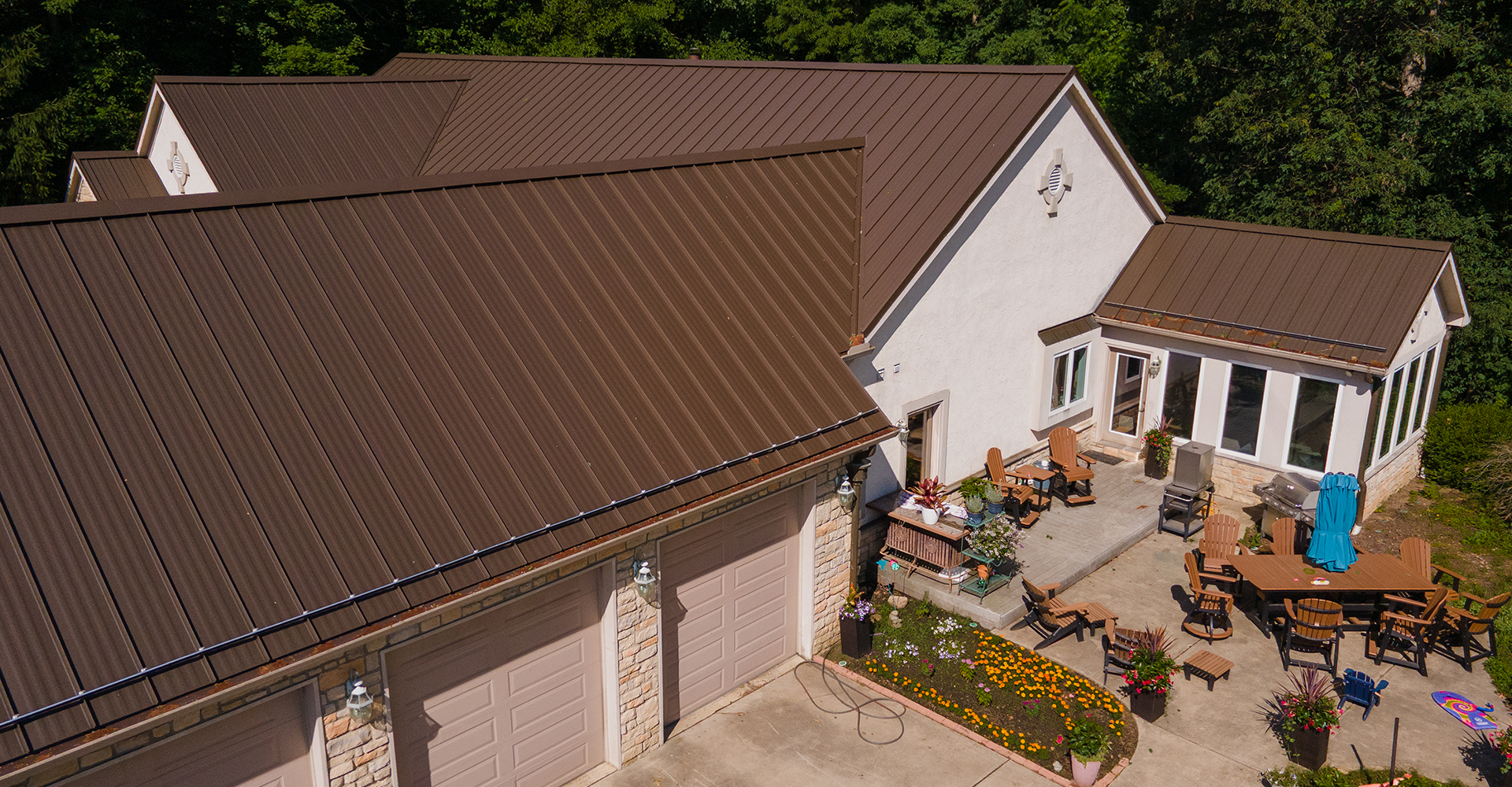 Aerial view of a house with a brown metal roof, three garage doors, a concrete patio with wooden chairs, tables, umbrellas, potted flowers, and a garden bed with orange flowers.