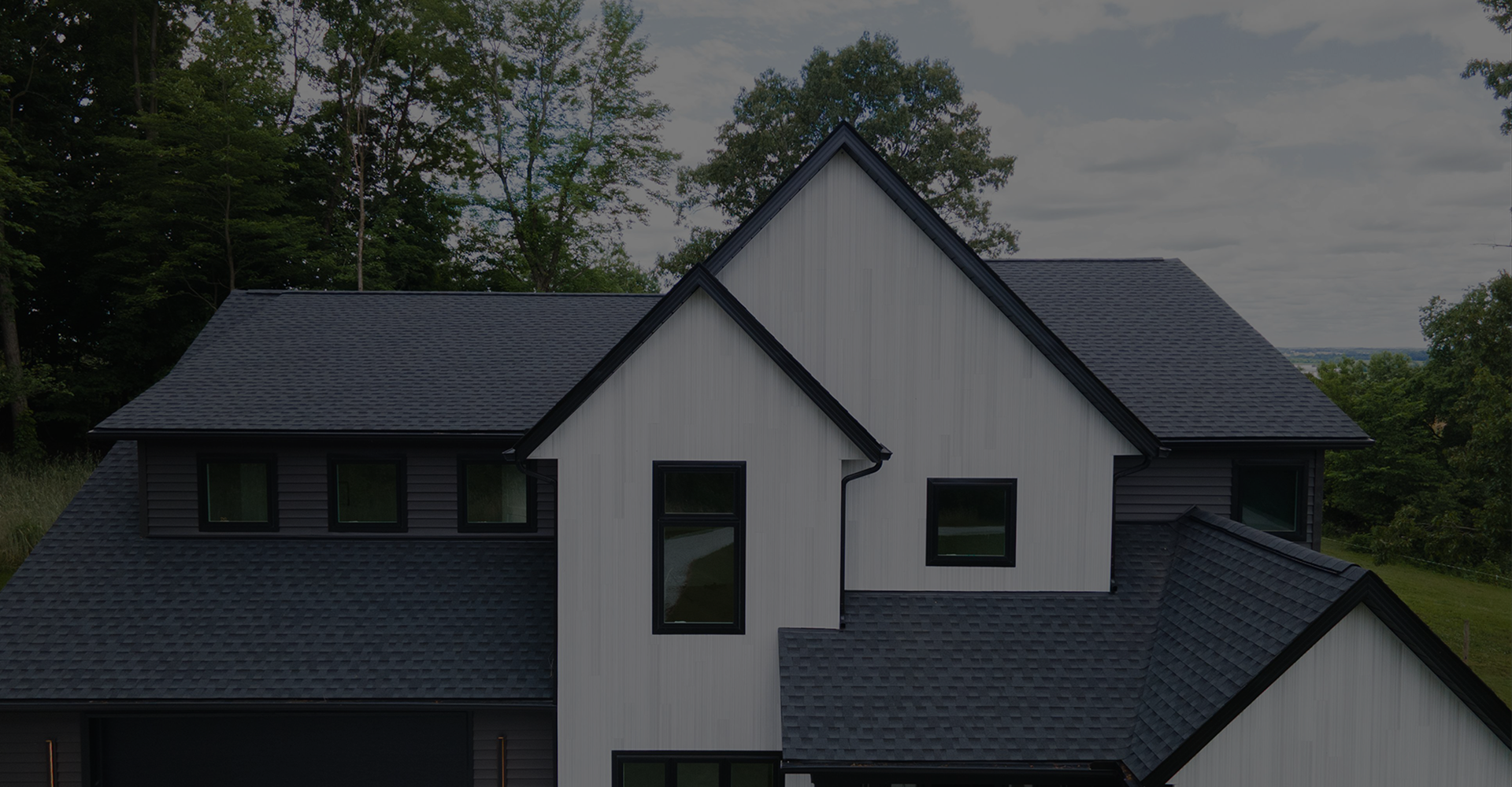 Modern house with dark gray shingle roofing and white siding accented by black window frames, surrounded by green trees.